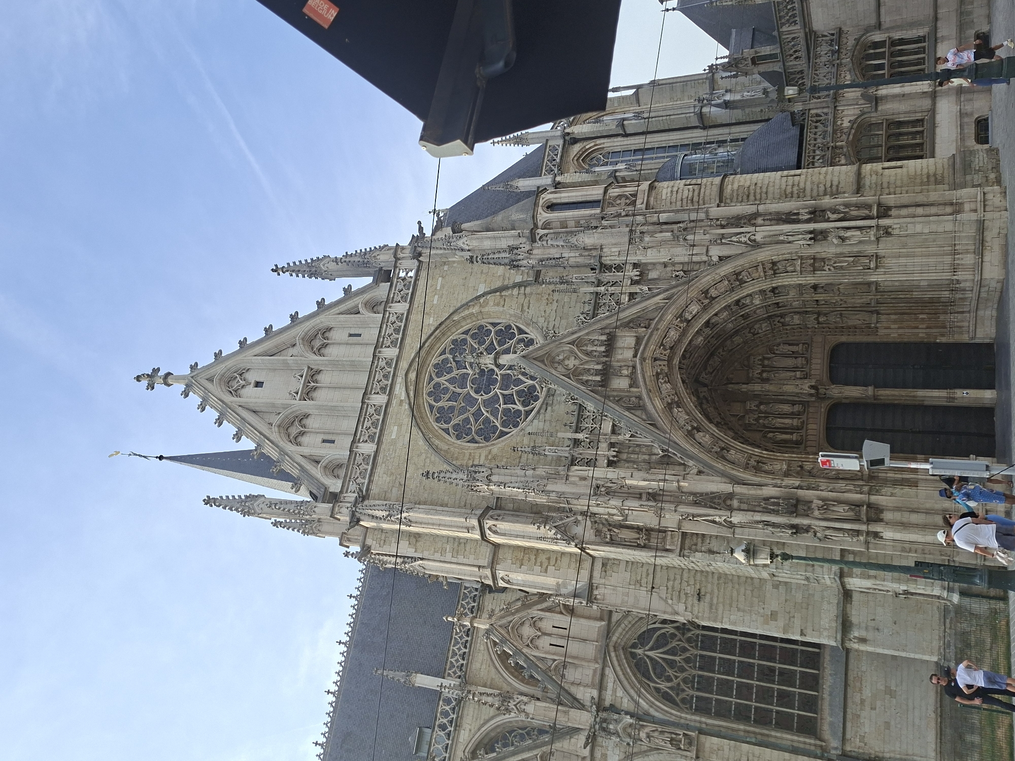 The image shows the facade of Saint Michael and Saint Gudula Cathedral in Brussels, Belgium, on a clear day. The cathedral is made of light-colored stone and features Gothic architecture with intricate carvings, pointed arches, and a large rose window above the main entrance. The main entrance consists of large, dark double doors. Several smaller windows with decorative tracery are visible on either side of the entrance. The cathedral is tall, with spires and pinnacles reaching towards the sky. A dark awning or canopy extends from the right side of the frame, partially obscuring the upper right portion of the cathedral. In front of the cathedral, several people are walking on a paved area, and a lamppost stands to the left of the entrance. The sky is a light blue with a faint cloud streak.