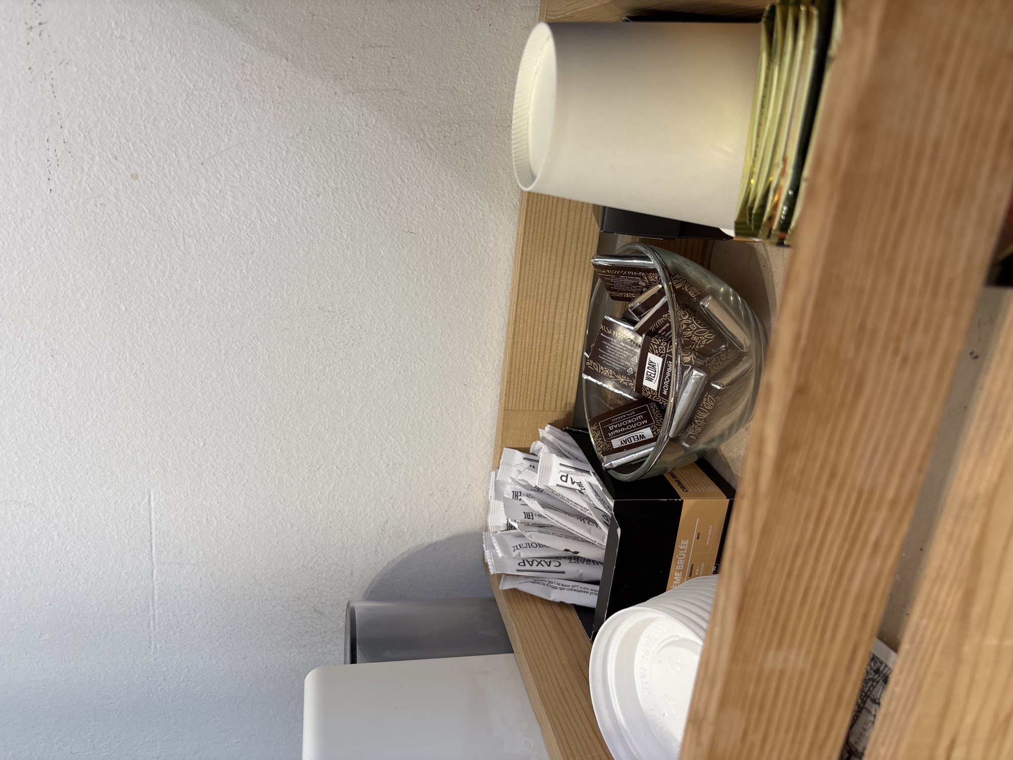 A wooden tray holds various items for making coffee or tea. In the back left, a white object and a grey cylindrical container are visible against a textured white wall. In the center of the tray, a black box is filled with individually wrapped sugar packets, some labeled "CAXAP". To the right of the sugar box, a glass bowl contains several individually wrapped chocolate squares, some with "WELDAY" visible on the packaging. In the front right, a white paper cup with a gold rim is positioned on the tray. In the front left, a white plastic lid is partially visible. The overall scene appears to be a coffee or tea station.