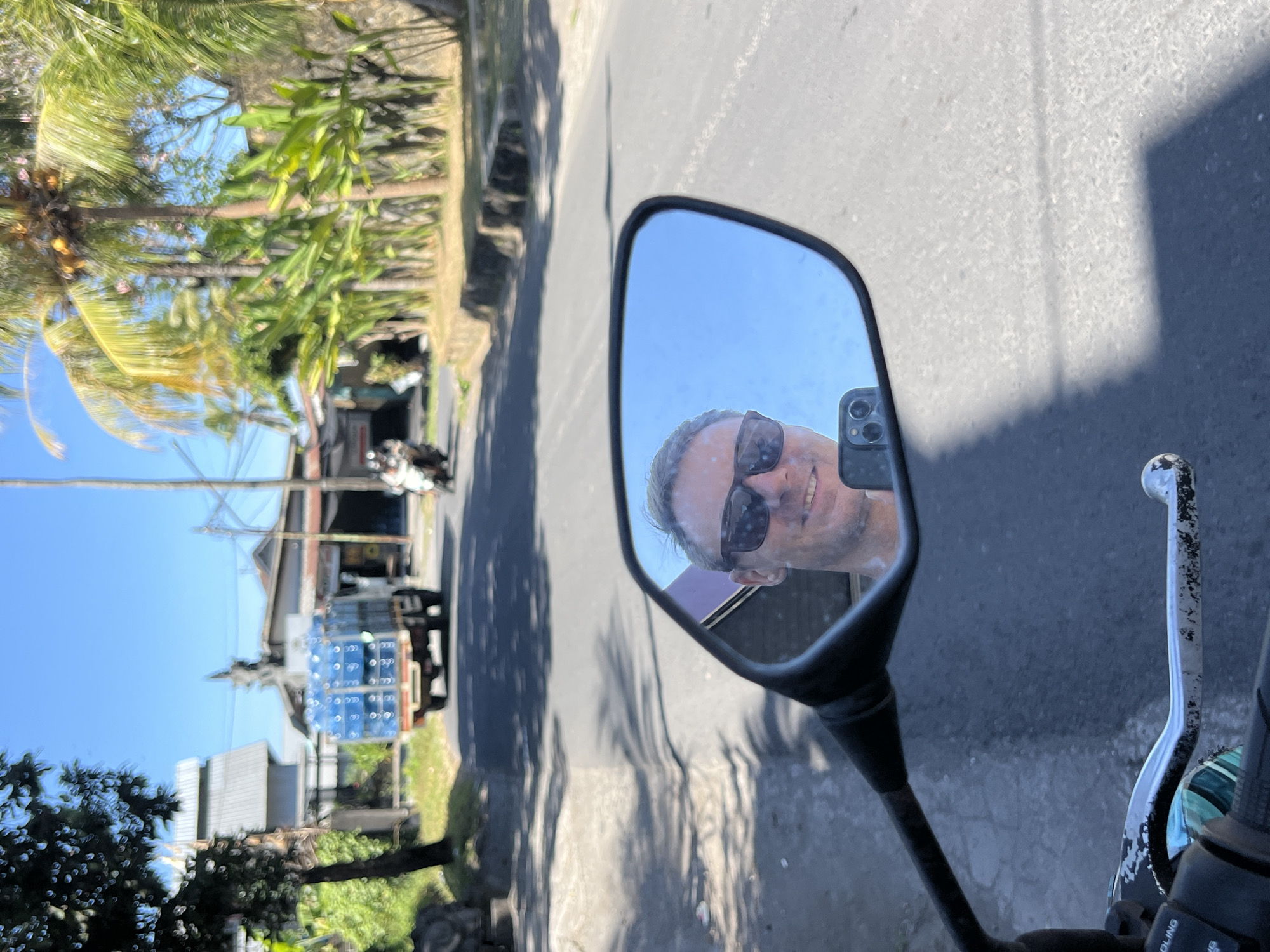 A man wearing sunglasses is reflected in the mirror of a motorcycle. He is holding a smartphone in his hand. The motorcycle is on a paved road in Sukawati, Indonesia, under a clear blue sky. In the background, a truck is carrying large blue containers, likely water bottles, and palm trees line the side of the road. The scene appears to be a sunny day in a tropical urban or semi-urban environment.