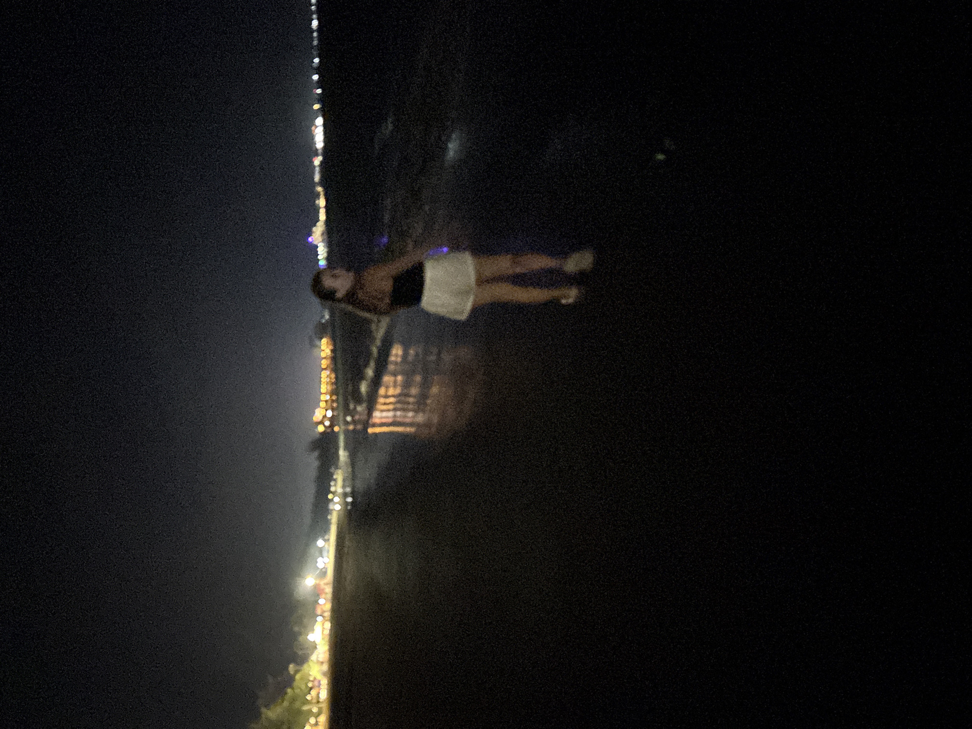 A grainy, low-light photograph captures a nighttime scene in Dong Hai Ward, Vietnam. The image is oriented sideways, featuring a single person, likely female, positioned towards the center. She appears to be standing in shallow water, dressed in a dark top and light-colored shorts or a skirt. Her body is slightly angled, and she is looking back over her shoulder towards the camera, suggesting a casual pose.
In the background, a distinct line of blurred, bright artificial lights stretches vertically across the frame, indicating a distant city skyline or well-lit shoreline. These lights cast subtle reflections onto the dark, calm water surrounding the subject. The sky above is uniformly dark, devoid of visible stars or moon, highlighting the urban illumination in the distance. The photograph's dark tones and visible grain are characteristic of a night shot taken in low light conditions. No discernible text is visible within the image.