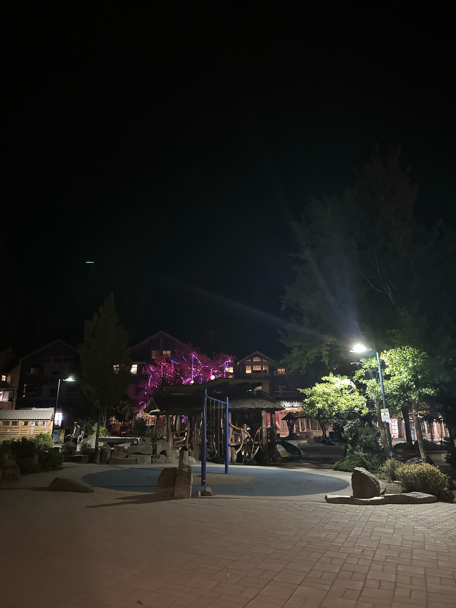 An outdoor nighttime scene shows a playground in the foreground with a paved brick walkway. A swingset with blue metal poles and a blue seat is visible. The playground area has a soft blue surface and is surrounded by large rocks. In the midground, several buildings with warm interior lights are visible, some adorned with bright pink and purple string lights. Trees with green leaves are scattered throughout the scene, illuminated by a bright streetlamp to the right. The sky above is dark.