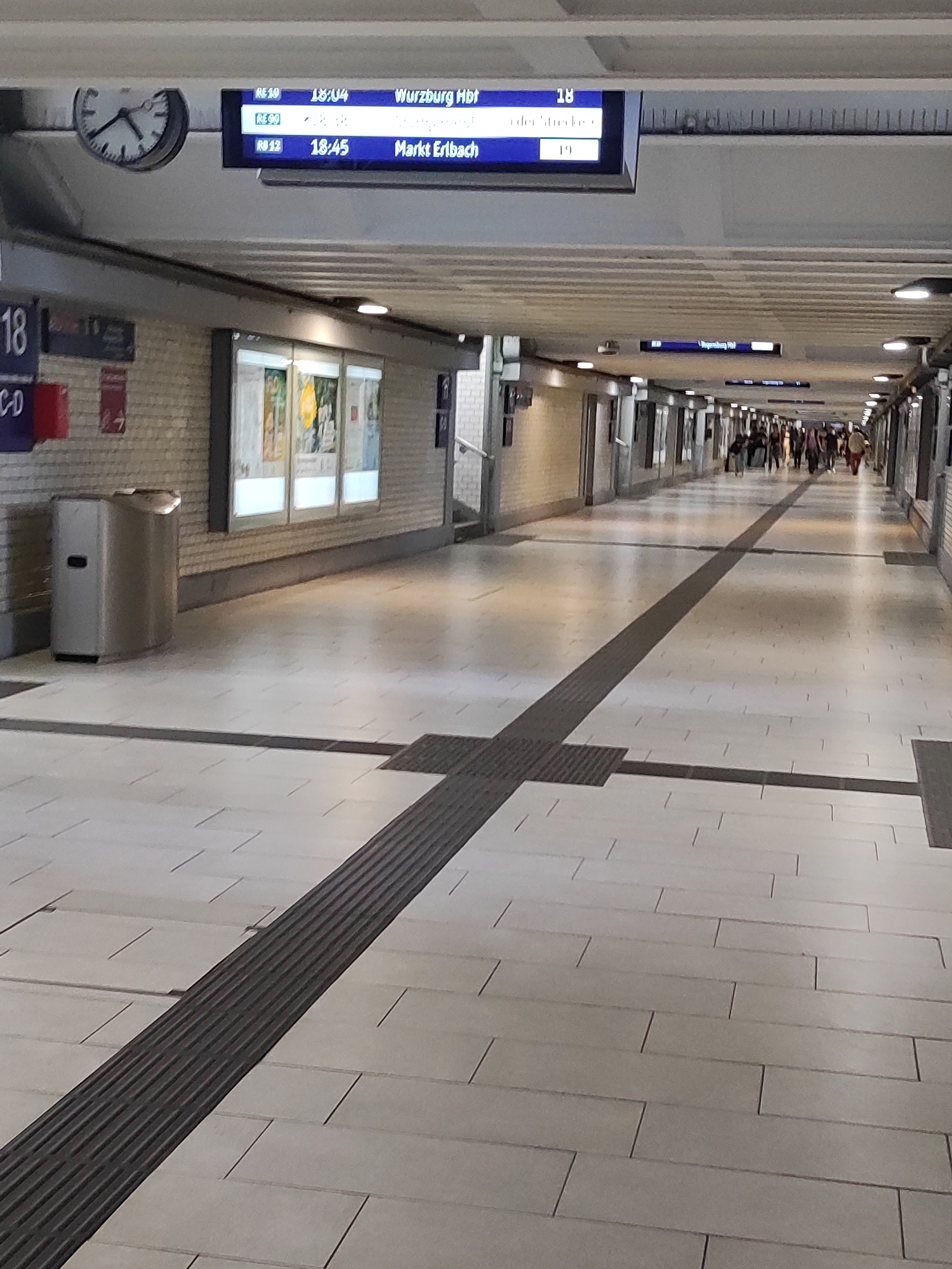 The image shows an underground pedestrian walkway, likely in a train station, given the signage displaying train schedules. The walkway is paved with light-colored tiles and features a dark, textured tactile paving strip running down the center, indicating a path for visually impaired individuals. The walls are lined with posters and light fixtures. In the background, a crowd of people is walking away from the camera. On the left side, a silver trash receptacle is visible, and above it, a digital display shows train information, including departure times and destinations such as "Würzburg Hbf" and "Markt Erlbach." A clock is also visible on the wall. The overall impression is of a functional, well-lit public transit space. Based on the presence of the train station signage and the architecture, this location is likely the Hauptbahnhof (main train station) in Nuremberg, Germany, given the typical appearance of German train station infrastructure and signage.