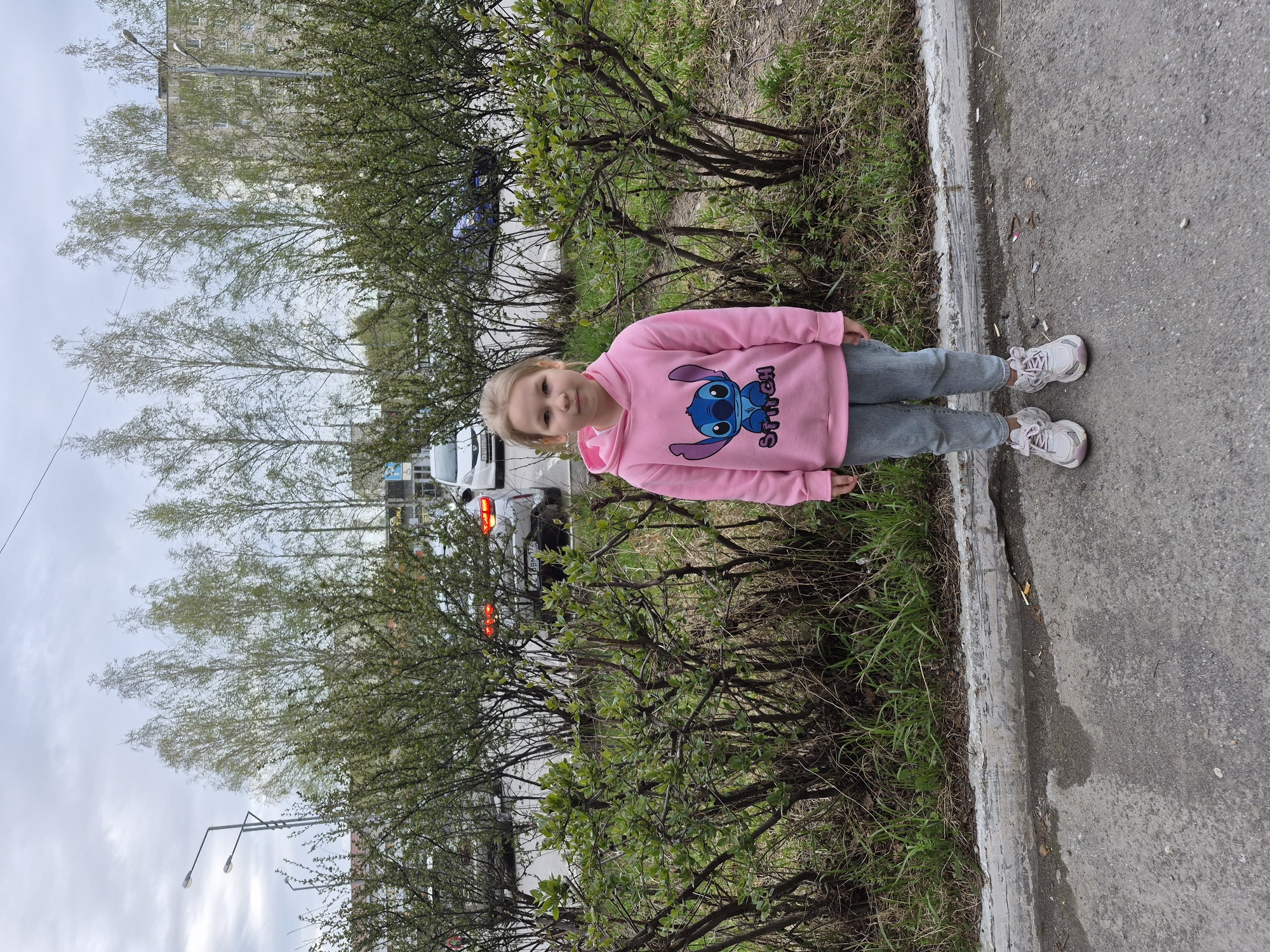 A young girl stands on a sidewalk next to a row of bushes. The girl is wearing a pink sweatshirt with a cartoon character on it, gray pants, and white sneakers. Behind the bushes is a street with cars and a building in the background. The location is identified as Tobolsk, Russia.