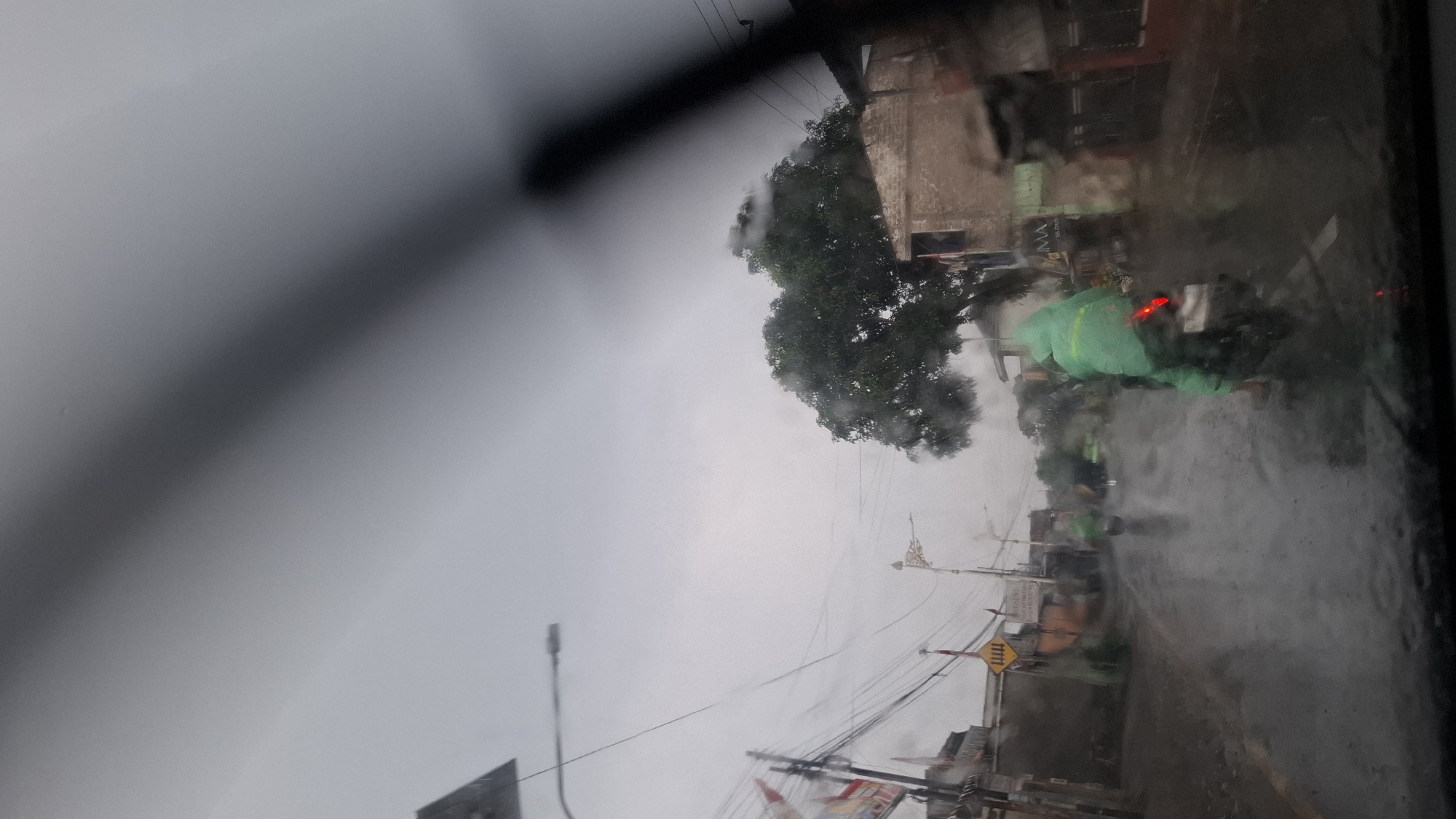The image depicts a street scene in Cilaku, Indonesia, during rainfall. The perspective is from inside a vehicle, looking through a rain-streaked windshield. Several motorcyclists are visible, most wearing bright green raincoats. One motorcyclist in the foreground is riding a motorcycle and appears to be wearing a green raincoat and pants, with their back to the camera. Other motorcyclists are further down the wet, reflective road. Buildings line the street, some appearing to be shops or houses. A large tree with dense foliage is situated on the right side of the frame, with its leaves also appearing wet. Utility poles and wires are visible above the street. The overall atmosphere is dim and overcast, consistent with rainy weather. The road surface is wet and reflects the surroundings.