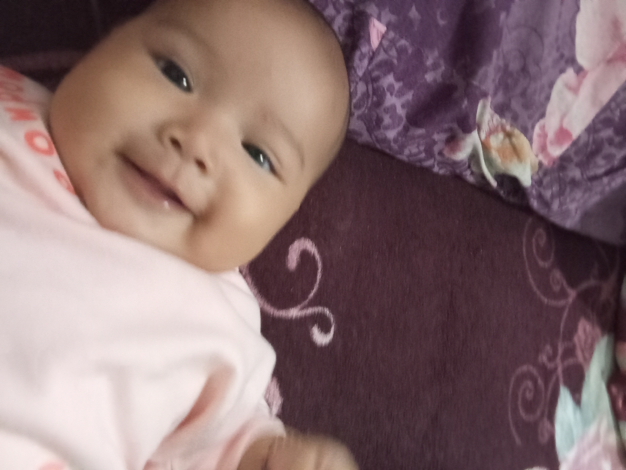 A close-up, low-angle shot captures a baby lying on a purple blanket with floral patterns. The baby, facing the camera, has a light brown complexion, dark eyes, and a wide smile. They are wearing a pink shirt with faint orange lettering. The background consists of the textured purple blanket, with a lighter purple patterned fabric on the left side and a darker, textured portion on the right with faint swirling designs. The image was taken indoors, likely in a residential setting in Rejosari, Indonesia.