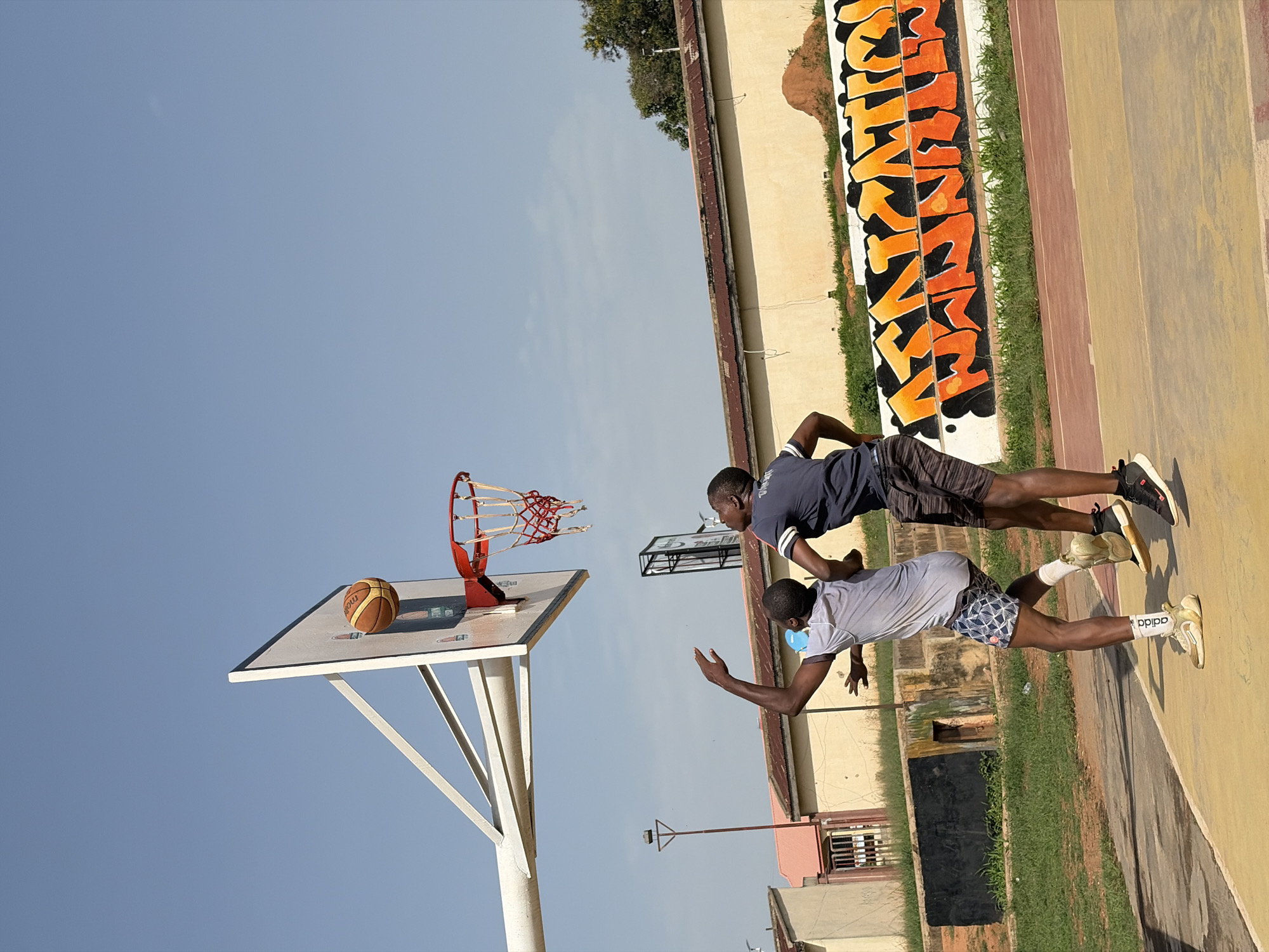 Two young men are on a basketball court in Jos, Nigeria. One is in a dark blue shirt and dark shorts, and the other is in a gray shirt, patterned shorts, a blue face mask, and white socks with Adidas sneakers. Both individuals appear to be in motion. A basketball rests on the backboard. The court is adjacent to a wall with graffiti, and the sky is blue.