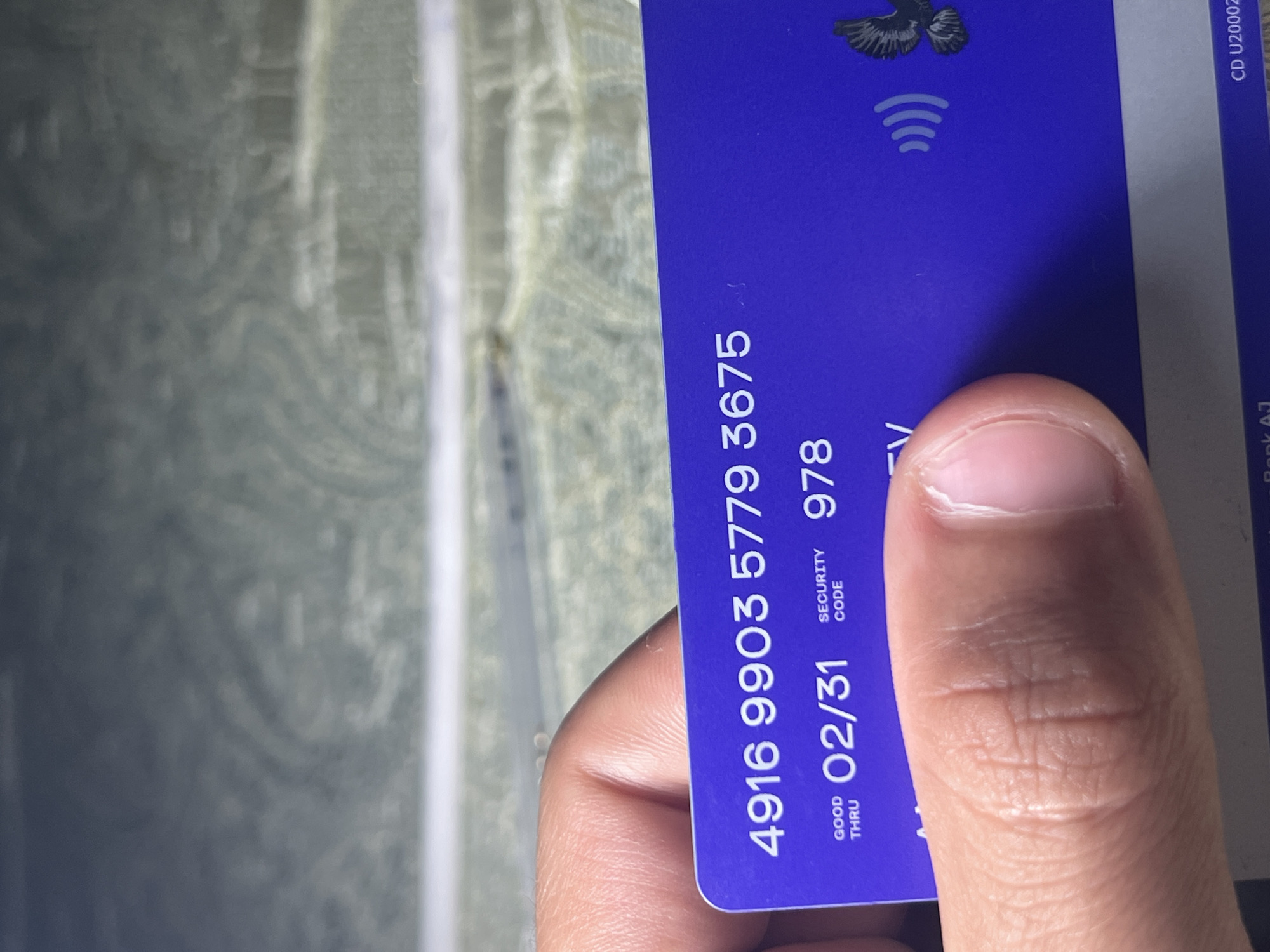 The image shows a close-up of a purple card being held by a person's thumb and index finger. The card displays alphanumeric text and a graphic of a bird with outstretched wings, as well as a contactless payment symbol. The visible text includes "4916 9903 5779 3675", "GOOD THRU 02/31", "SECURITY CODE 978", and "TV". The background of the image is out of focus and appears to be a textured surface with a pattern of swirling lines, possibly resembling wood grain or marble. The lighting suggests the image was taken indoors or in an area with ambient light. The location is identified as Romitan, Uzbekistan.