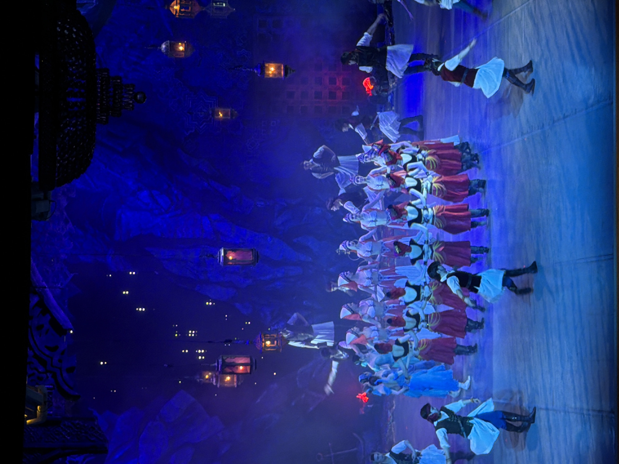 The image shows a stage performance in Izhevsk, Russia. A large group of performers, mostly women, are arranged in rows, wearing costumes consisting of white headwear, black vests, white shirts, and red or blue skirts. Some performers are jumping. Lanterns hang from the stage ceiling. The backdrop is blue.
