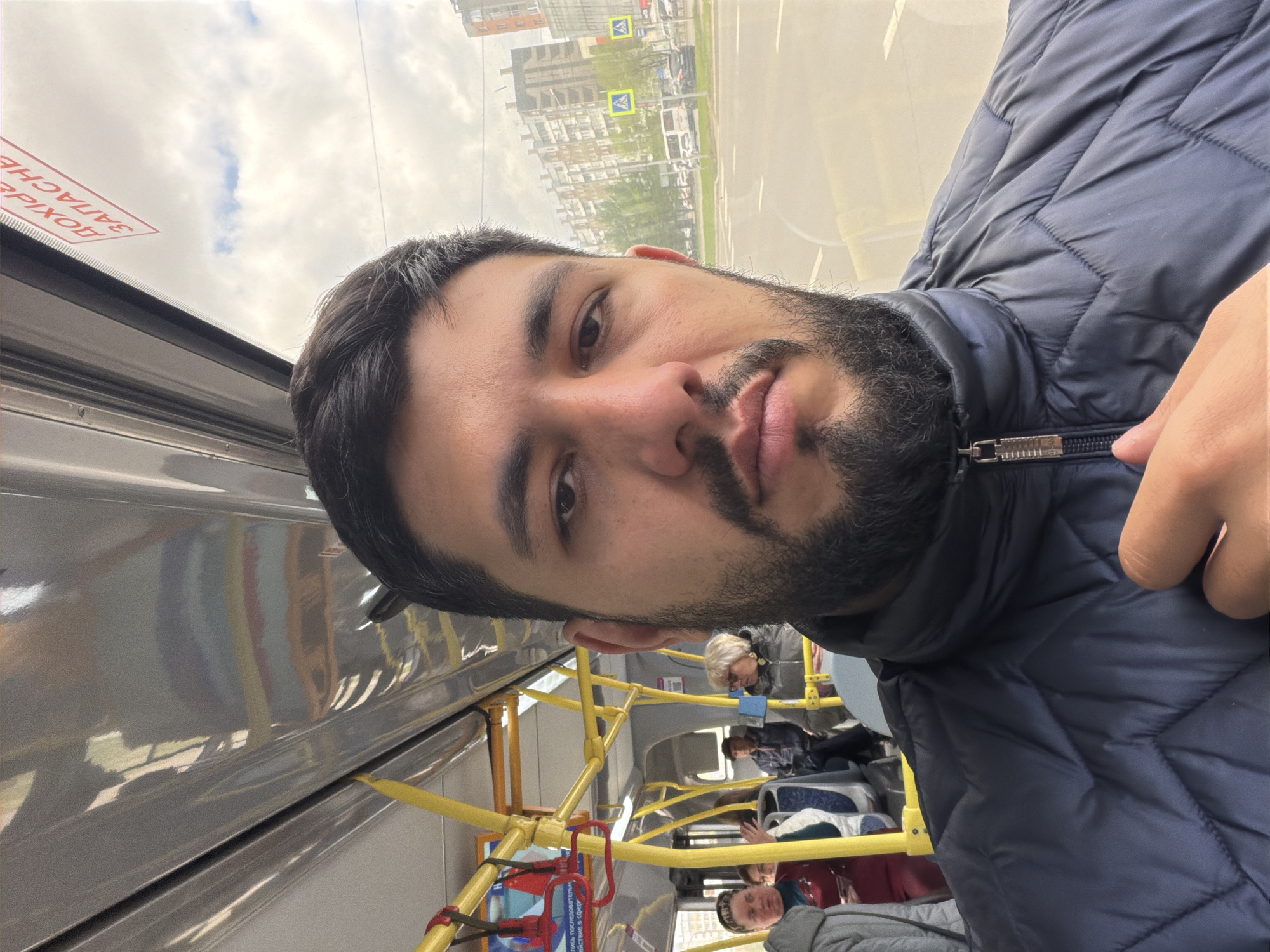 The image depicts a man with dark hair and a beard inside a public transportation vehicle, likely a bus, in Krasnoyarsk, Russia. The man is wearing a blue puffer jacket. Passengers are seated in the background. Through the window, buildings and street signs, including the yellow pedestrian crossing signs commonly used in Russia, are visible. The interior of the bus features yellow handrails.