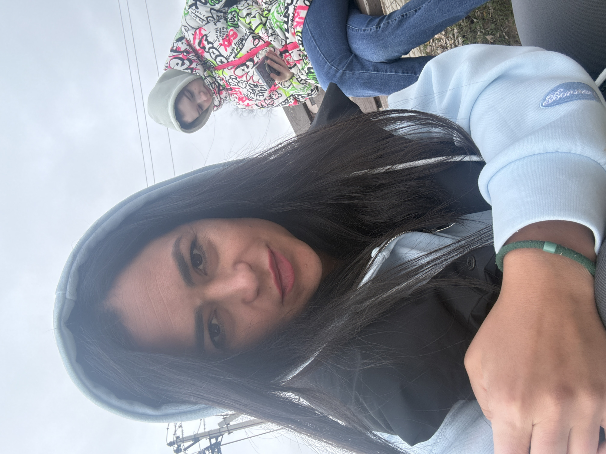 The image, captured from a low angle, features two individuals outdoors on an overcast day in Chaikovsky, Russia. The person in the foreground, taking a selfie, has long dark hair flowing down and wears a light blue hooded sweatshirt over a dark zippered jacket or vest. A green beaded bracelet is visible on her left wrist. She looks directly at the camera with a subtle smile and reddish lips, her dark eyes prominent. An embroidered logo on her left sleeve appears to spell "Bonjour" in cursive script.
Behind her, another person is seated on a wooden bench. This individual is wearing a distinctive hooded jacket with a white base adorned with colorful graffiti-style text and patterns in pink, green, and black, including words such as "Negative," "Positive," "Happy," and "Crazy." She is dressed in blue jeans and holds a dark smartphone in her hands, looking towards the camera with a neutral expression. Her hood is up, framing her face.
The setting is outdoors, with a uniformly grey and overcast sky filling a significant portion of the background, indicating a cloudy daytime. In the distant background, power lines or a utility pole structure are visible, suggesting an urban or semi-urban environment. The overall lighting is soft and diffused, characteristic of an overcast day, and there are hints of bare tree branches in the background, implying late winter or early spring. Both individuals appear to be casually dressed, possibly enjoying a relaxed moment outdoors.
