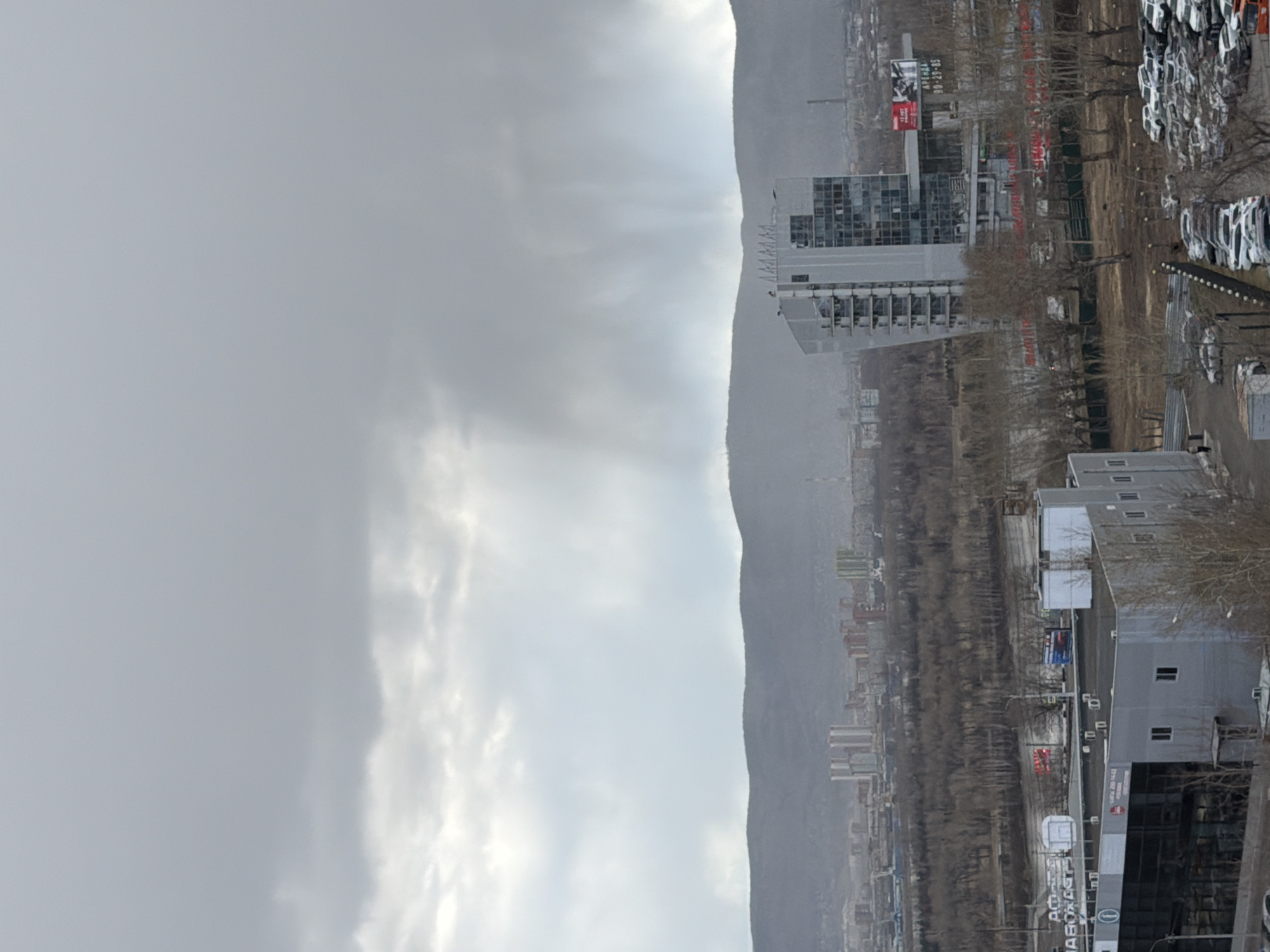 The image presents a broad view of an urban landscape set against a mountainous backdrop under an overcast sky. The prominent feature in the background is a long, undulating mountain range, appearing barren or sparsely vegetated, stretching across the horizon. Above it, the sky is largely gray and cloudy, with hints of brighter light breaking through the clouds, creating faint crepuscular rays that pierce downwards.
In the mid-ground and foreground, an urban environment unfolds. To the right, a vast parking lot is densely packed with numerous white cars, suggesting a car dealership or a storage area for new vehicles. Scattered bare trees, indicative of winter or early spring, line parts of the landscape. Several buildings are visible, including a modern, multi-story structure with glass and gray facades in the right foreground. Further back, another taller building with numerous windows and visible balconies or architectural details rises amidst other structures. In the distance, closer to the mountain base, various residential or industrial buildings are discernible.
There is no specific human activity evident, presenting a static city scene. The overcast weather and the barren appearance of the trees and mountains contribute to a subdued, somewhat cool atmosphere.
Several pieces of text are visible on signs and buildings. On a billboard in the upper right, "АВЕНЮ 90-29-45" is displayed. A prominent sign on the building in the foreground right clearly reads "АВТОПАРКЪ" (AUTOPARK) along with a phone number such as "589-780-14-23" and partially visible text "ТОРГОВЫЙ" (TRADE). Another sign on a pole has the warning "НЕ ПОДХОДИ БЛИЗКО" (DO NOT COME CLOSE). The presence of Russian text, the urban development, and the distinct mountain range are consistent with the provided location context of Krasnoyarsk, Russia.