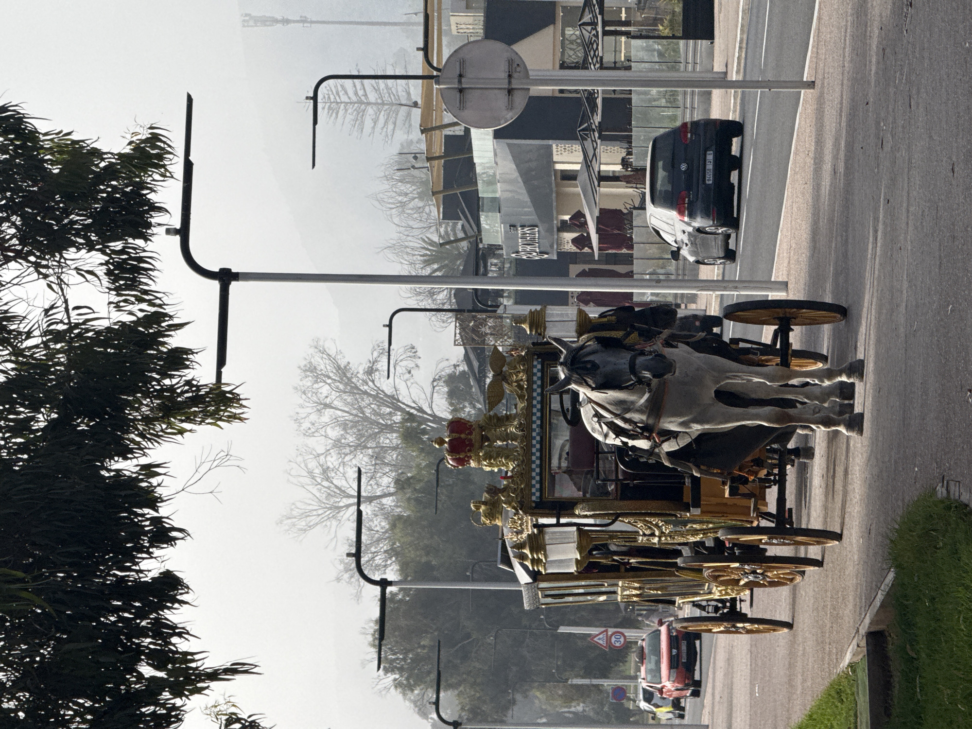On a misty day in Agadir, Morocco, a horse-drawn carriage occupies the right-hand lane of a paved road. The carriage is elaborately decorated in gold and bears a decorative red crown. It is being pulled by two white horses. The carriage has multiple wooden wheels. The traffic lane is also occupied by parked cars. Street lights line the road. Buildings and trees are visible in the background. The license plate "84458-131 6" is visible on a dark-colored car.