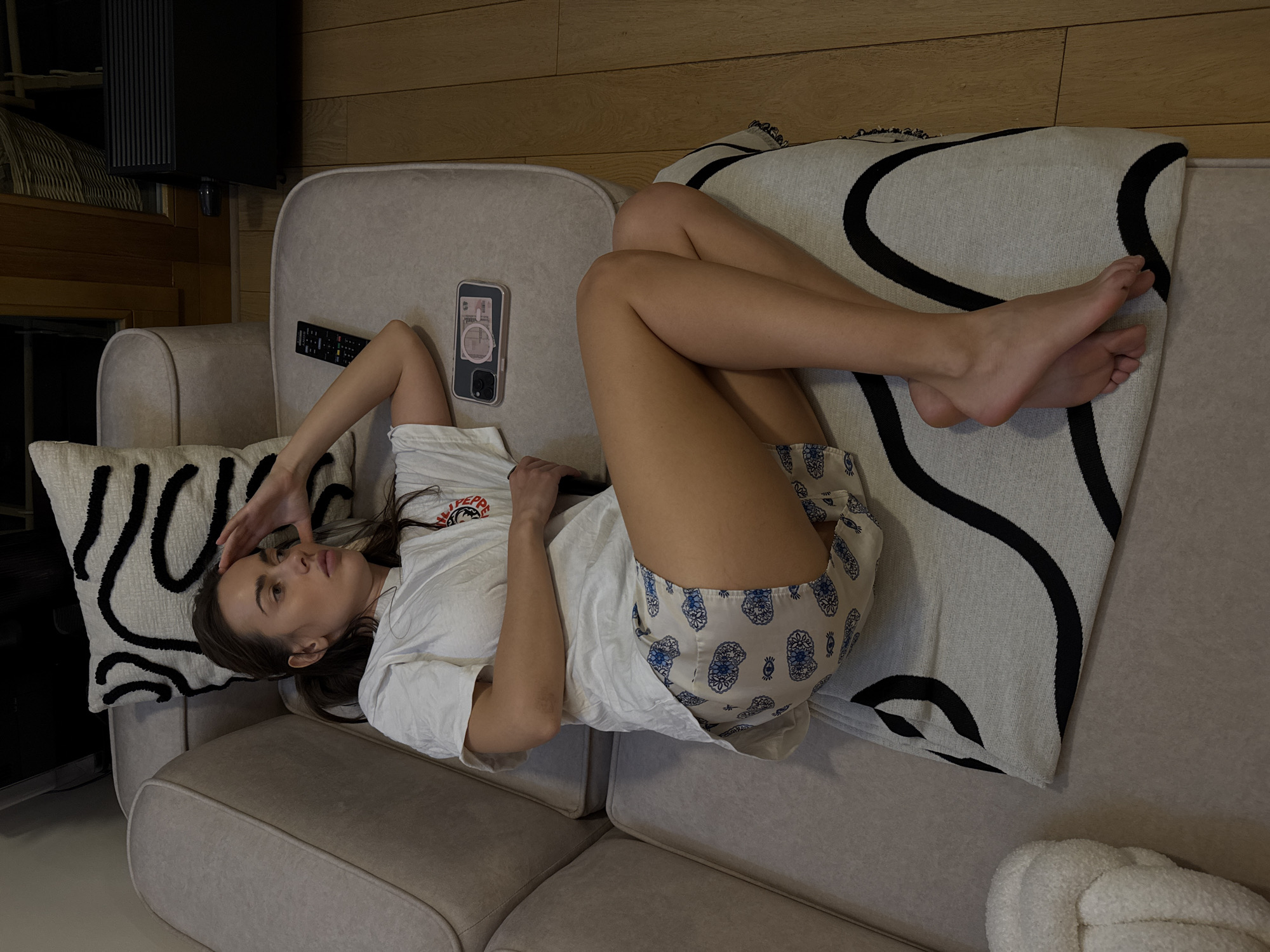 A woman is lying on a beige couch. She is wearing a white t-shirt and patterned shorts. Her right arm is raised, and her hand is touching her forehead. Her legs are crossed near the couch's end, with bare feet visible. Beside her left hand is a remote control and a cellphone with a pink design on the case. A black and white patterned pillow is behind her head. The couch has a similar patterned throw at the opposite end where her feet rest. The background shows a wooden wall.