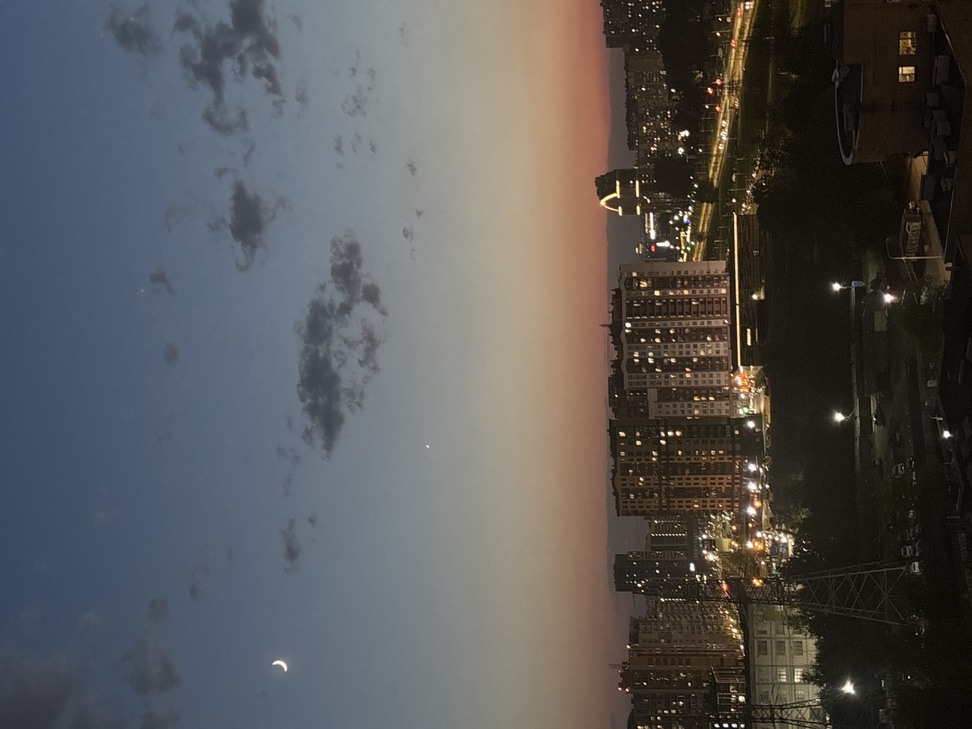 Here's a detailed description of the image:
The image presents an evening cityscape under a twilight sky. The sky transitions from a deep blue with scattered clouds on the left side, to a gradient of orange and gray near the horizon on the right. A crescent moon is visible in the lower left portion of the sky. A small, bright celestial object, possibly a star or planet, is also visible in the center.
The urban landscape is characterized by high-rise buildings densely packed together. Their windows are lit, indicating residential or commercial activity. The buildings are silhouetted against the orange sky. At street level, lights from cars and street lamps illuminate the roads and surrounding areas. The urban area includes highways and green spaces. There is a telecommunications tower at the bottom of the frame.