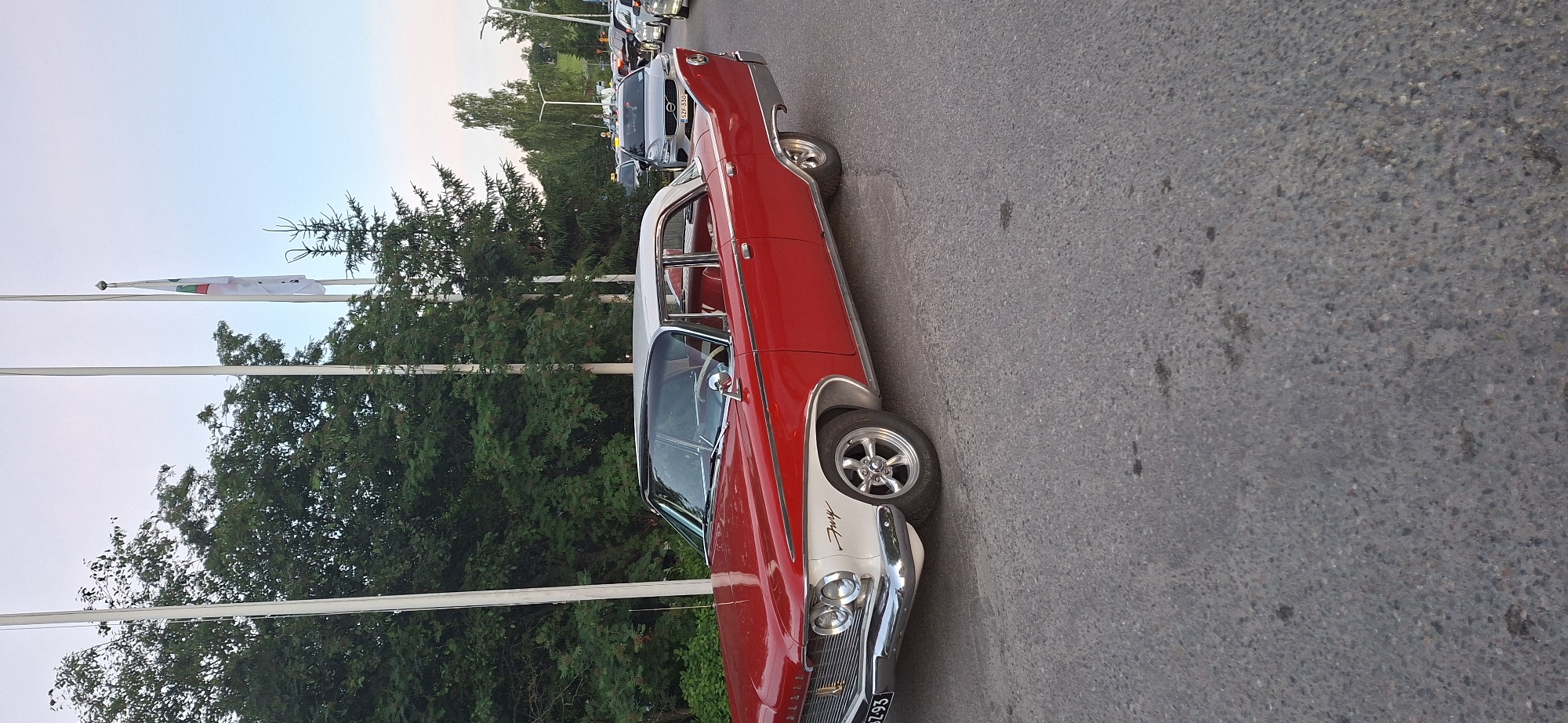 The image depicts a classic red and white two-door sedan, identified as a 1957 Plymouth Fury, parked on an asphalt road. The car is the primary subject, positioned slightly to the left of the frame, facing right. Its distinctive chrome grille, dual headlights, and iconic tail fins are visible. A license plate with the characters "SZ-93" is affixed to the front. The car's interior, featuring red seats, is partially visible through the windows.
In the background, several other vehicles are present, including a silver Volvo sedan and what appears to be a taxi. Trees and flagpoles line the right side of the road. One flagpole displays a flag with green, white, and red vertical stripes and a symbol. The environment suggests an outdoor setting in a town or city, possibly during daylight hours with overcast skies. The image was taken in the city of Salpakangas, Finland.