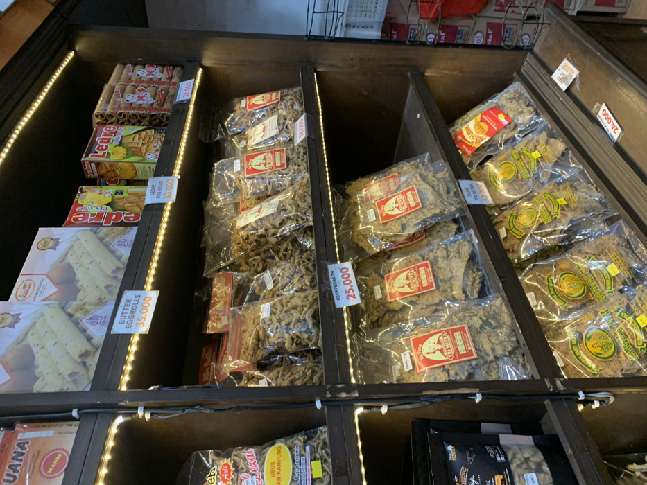 A wooden shelf unit with four shelves in a shop located at latitude -7.584, longitude 110.287 in Muntilan, Indonesia, is stocked with various packaged snacks. On the top shelf, there are three packages of snacks with price tags: "BUTTER EGGROLLS 35.000," "LEDES DUS HELLO 30.000," and "LEDES 32.000." On the second shelf, there are numerous transparent bags filled with crispy fried snacks, with one price tag: "USUS PEDAS TM 25.000." On the third shelf, there are transparent bags filled with a different type of crispy fried snack, with one price tag: "PARU TRITIMA 67.000." On the bottom shelf, there are several transparent bags filled with a different type of crispy fried snack, with one price tag: "26.000." There is a bright white light strip running along the top of the shelf unit. The shelves appear to be made of dark wood. The background behind the shelves is out of focus and partially obscured. There is a bright red item in the background on the right side of the image. The image is focused on the shelves and the snacks for sale.