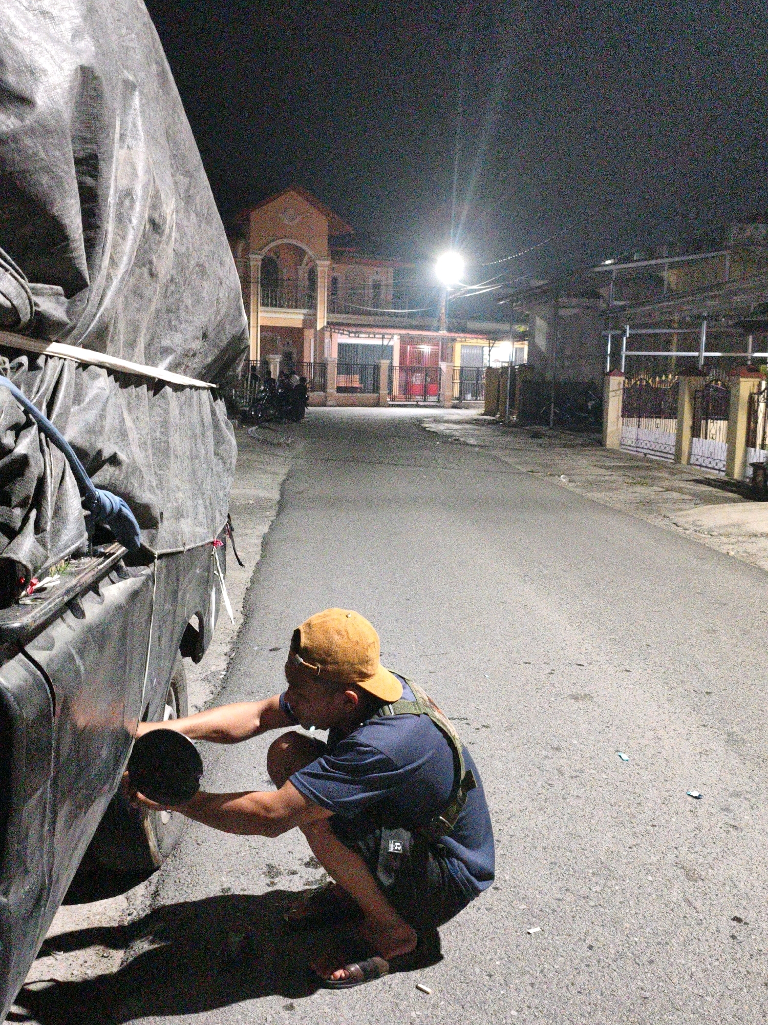 In Koto Baru Semurup, Indonesia, at night, a person wearing a blue shirt, dark shorts, and an orange baseball cap squats by the side of a black truck. The truck is covered with a large, gray tarp. The individual is holding a black, round object near the truck's wheel well, possibly engaging in maintenance or inspection. A bright street light illuminates the scene, casting a long shadow of the person onto the asphalt road. In the background, houses with illuminated windows line the street, and a few motorcycles are parked. The environment appears to be a residential street at night.