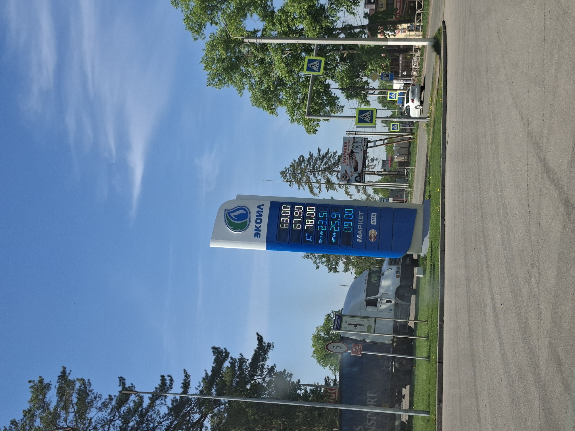 The image captures a gas station under a bright, clear sky. The prominent sign displays the station's brand, "ЭКОИЛ," along with fuel prices: 63.00, 67.90, ДТ at 78.00, ПРОПАН at 23.50, МЕТАН at 25.90, and 09.00. Payment options include Mastercard and VISA.
Behind the sign, a semi-truck is visible, along with a sign with the number "5" on it. The background includes trees and a glimpse of buildings. Additionally, a sign for "ЗАПРАВКА АВТОКОНДИЦИОНЕРОВ" is present. A white car is seen farther up the road, past multiple pedestrian crossing signs. The city is Unknown, Unknown.