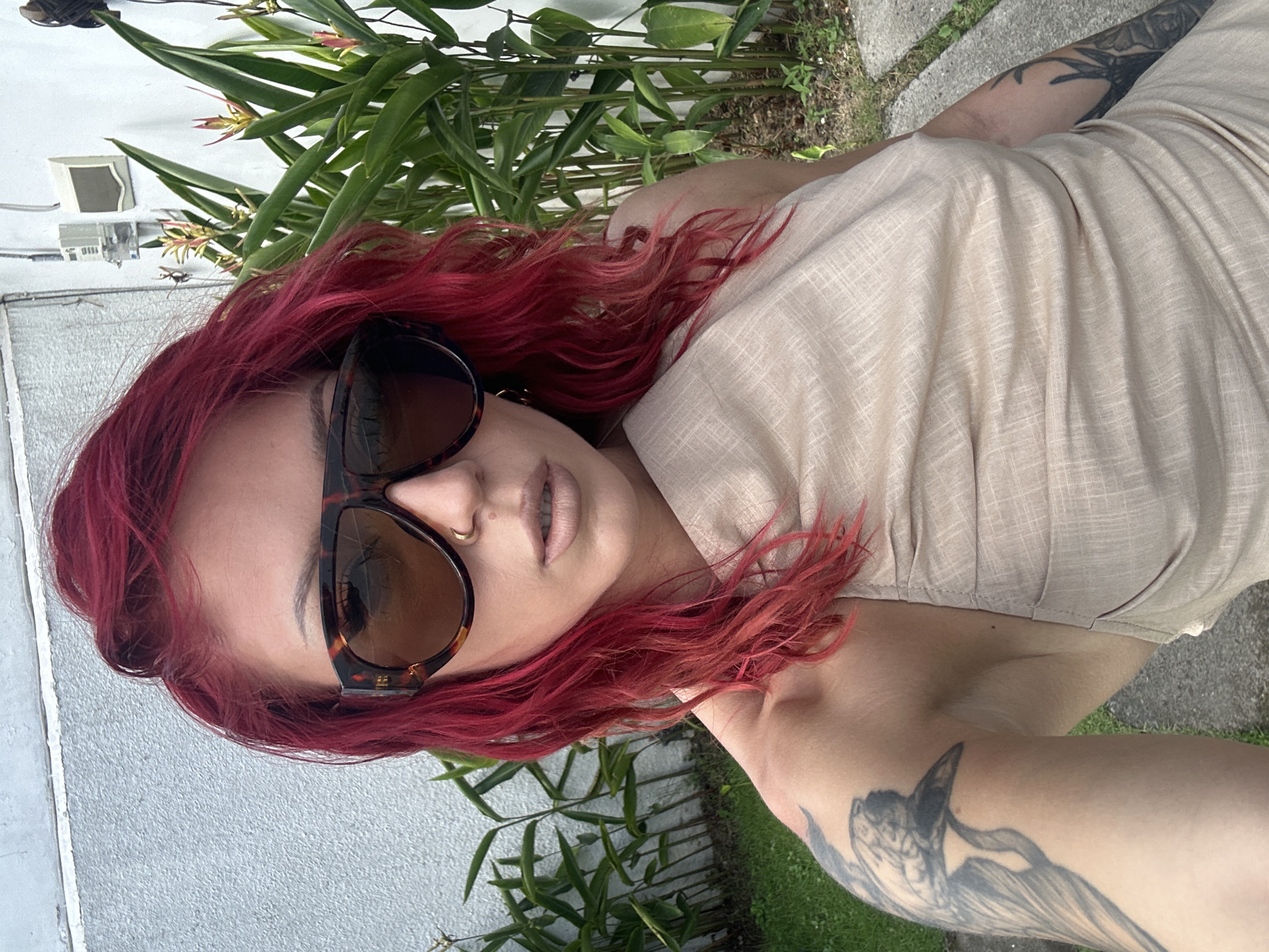 A woman with red hair and sunglasses stands outside near foliage, possibly in North Kuta, Indonesia. She is wearing a tan top and has tattoos on her arms. The background includes a white wall and greenery.