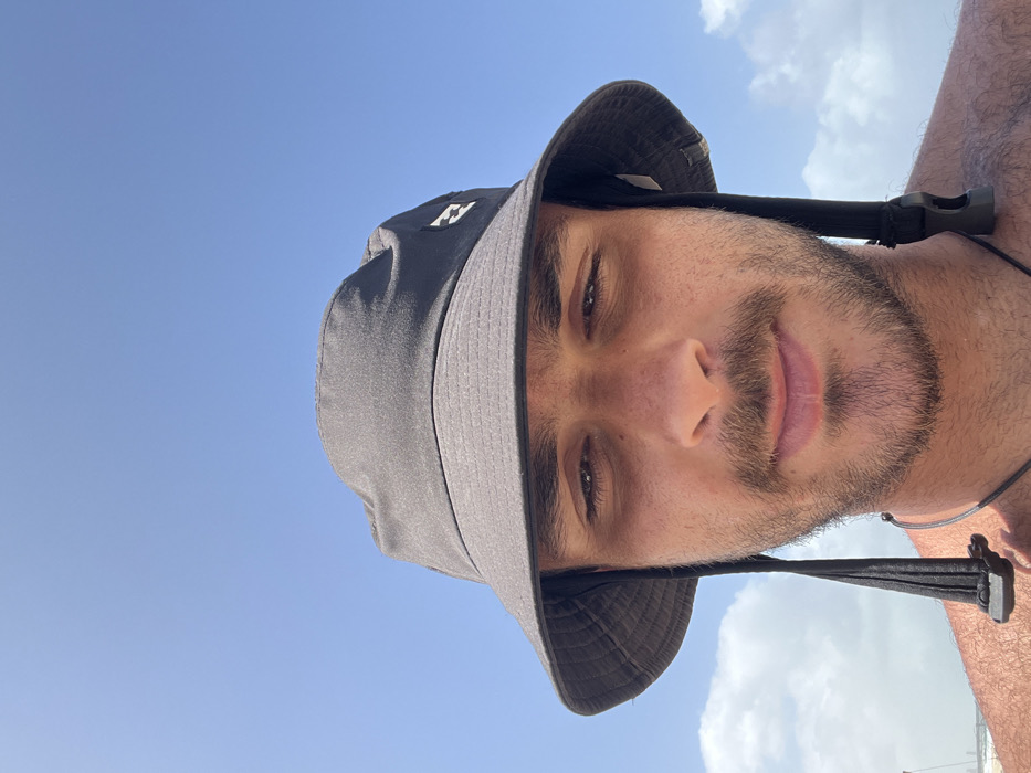 The image is a close-up selfie of a man wearing a black bucket hat with a small white logo on the side. He has short brown hair, a beard, and a mustache. His eyes are open, and he is looking directly at the camera. His expression is neutral. His torso is visible, as well as part of his bare chest and arms. He is wearing a black necklace. The background is a blue sky with some clouds. The image appears to have been taken on a beach in Sdot Yam, Israel, as indicated by the coordinates provided.