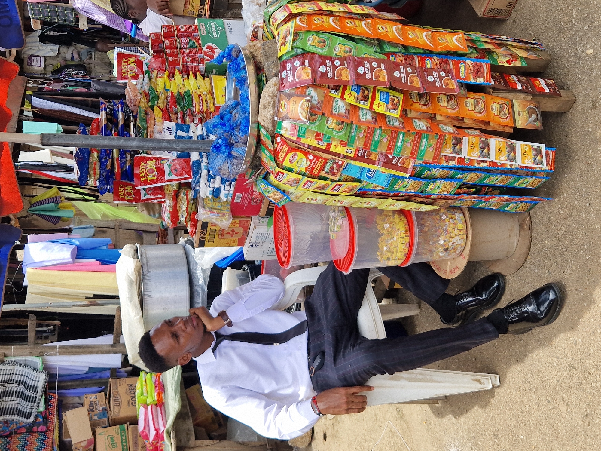 A young Black man wearing a white dress shirt, black tie, and pinstriped pants sits on a white plastic chair facing left, with his left hand resting on his chin. He is in an outdoor market setting. Behind him, to his right, is a stall with an assortment of food products, including bags of seasoning and rice mixes hanging from a wooden frame. Red-lidded plastic containers filled with yellow cubes are stacked on a wooden spool to his right. In the background, various goods are displayed, including fabric, boxed products, and large metal pots. The ground is a paved surface. The overall scene depicts a market stall with a customer or vendor present.