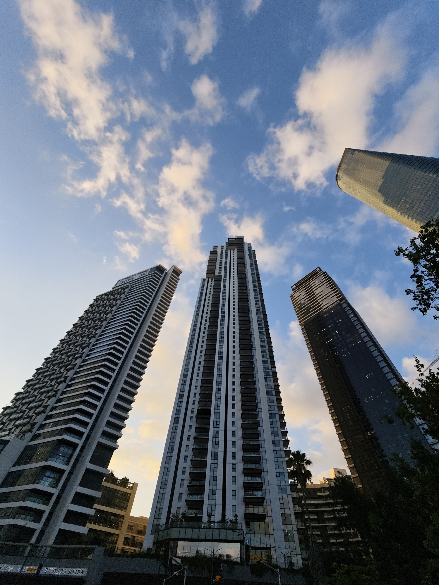 The image captures a low-angle view of several modern skyscrapers against a sky filled with scattered clouds and a hint of sunset colors. In the foreground, three tall buildings are prominently featured. The leftmost building is characterized by its repetitive, angular facade with numerous balconies. The central skyscraper is the tallest, with a sleek, multi-faceted design, showcasing a complex arrangement of windows and balconies. To the right, a darker, glass-clad tower rises, reflecting the sky. In the upper right, a portion of a rounded, tower-like structure is visible, possibly a cooling tower or another type of cylindrical building. The sky is a blend of blue, with patches of yellow and orange from the setting sun illuminating the clouds. Trees and other vegetation are visible in the lower right and between some of the buildings, suggesting an urban park or landscaping. The scene appears to be set in a contemporary urban environment, likely a business or residential district. Based on the Hebrew text visible on a sign in the lower left corner ("אחוזה הבנאים"), which translates to "Builders' Estate", and the architectural style, the location is identified as Tel Aviv, Israel. No people or specific actions are discernible in the image.
