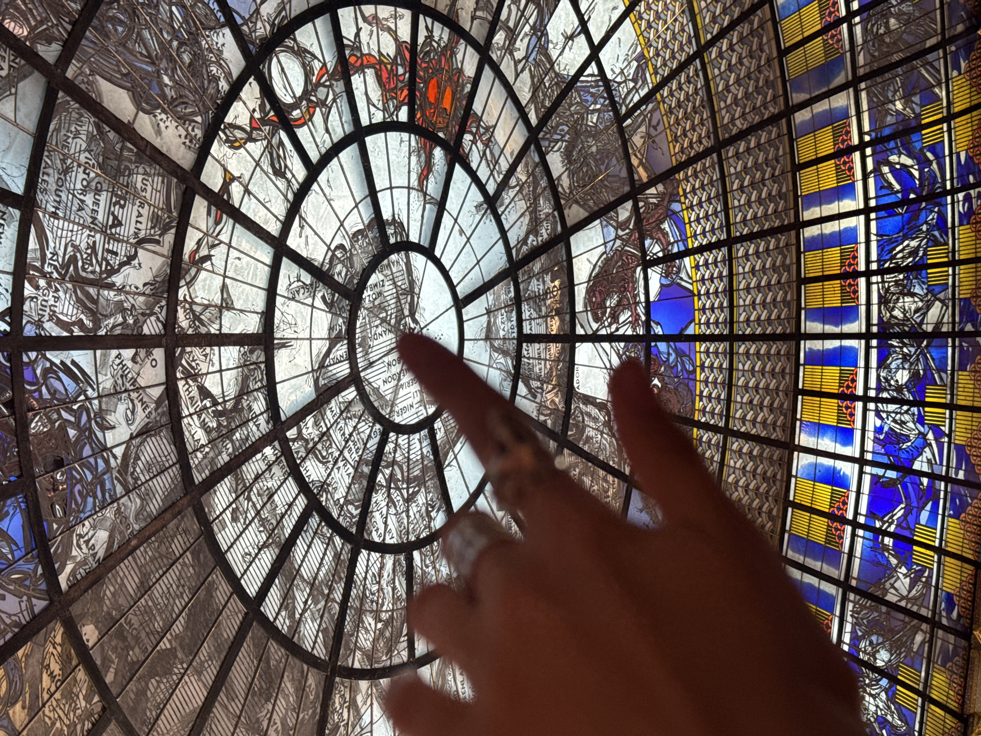 A hand with rings on its fingers is reaching up towards a stained-glass dome ceiling. The dome is intricate and features sections with maps and illustrations, including depictions of animals and geographical locations. The lower part of the dome has bands of yellow and blue stained glass. The setting appears to be indoors, possibly a public building or museum, within Samrong Nuea Subdistrict Municipality, Thailand.