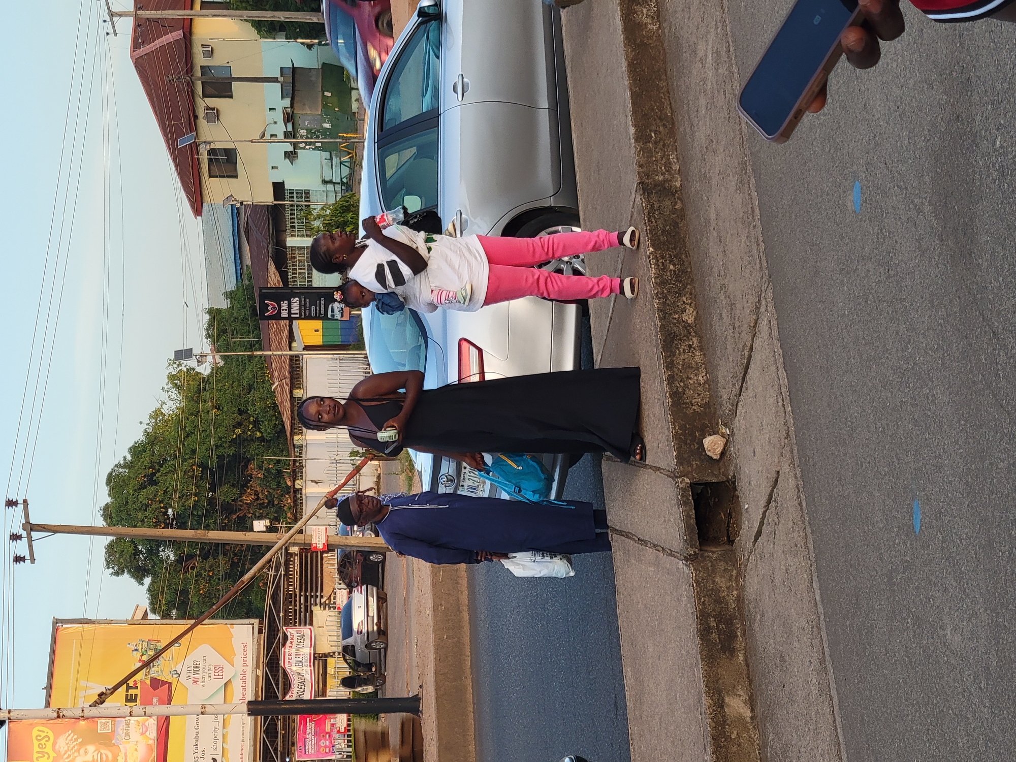 Here is a description of the scene:
Three individuals stand on a concrete sidewalk adjacent to a city street. To the right, a person holds a mobile phone, partially visible. The next individual is wearing a blue robe with a white bag. Then next person is wearing a black dress. The fourth person is holding a child, and is wearing pink pants and a white shirt. A silver car is parked next to them. The scene is on a city street in Jos, Nigeria, as evidenced by the street sign with "35 Yakubu Gowon Way, Jos." The sign includes text promoting the shopcity Jos supermarket. Additional signage in the scene includes "Deng Links." Trees, buildings, and utility poles are visible in the background.