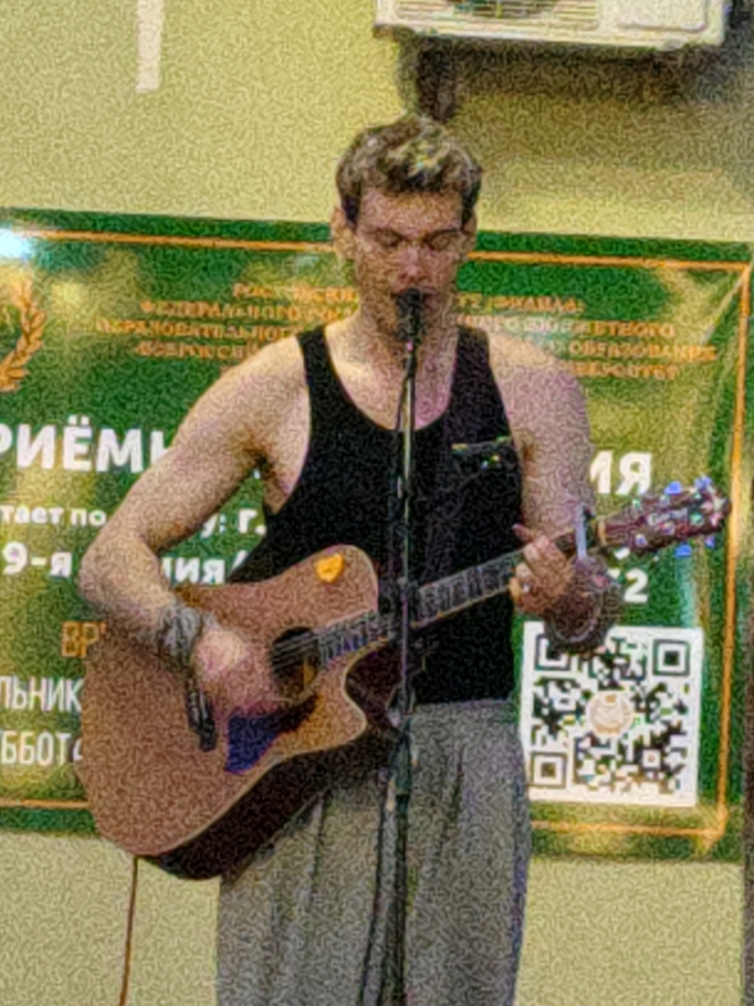 A young Caucasian male with short, wavy brown hair and glasses is performing with an acoustic guitar in front of a banner in Rostov-on-Don, Russia. He is wearing a black tank top and grey pants. He is holding the guitar with his left hand and strumming with his right. He is looking down at his guitar. The banner behind him is green with white lettering. There are also two QR codes visible on the banner. The lighting is dim and the image is grainy.