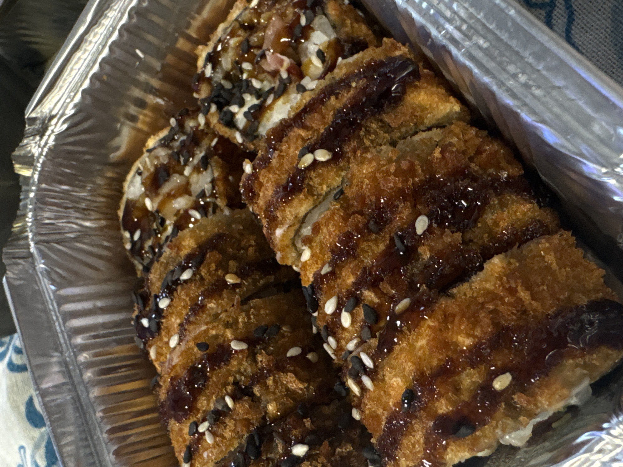 A close-up, overhead shot shows a foil container filled with several pieces of deep-fried sushi rolls. The sushi is coated in a golden-brown breading and drizzled with a dark, glossy sauce, likely teriyaki or eel sauce. Black and white sesame seeds are sprinkled on top. The rolls appear to be cut into bite-sized pieces, revealing a white interior, possibly rice and a filling. The container is placed on a textured surface, possibly a tablecloth with a blue and white pattern, within the city of Yarominski Sel'ski Sovet, Belarus.