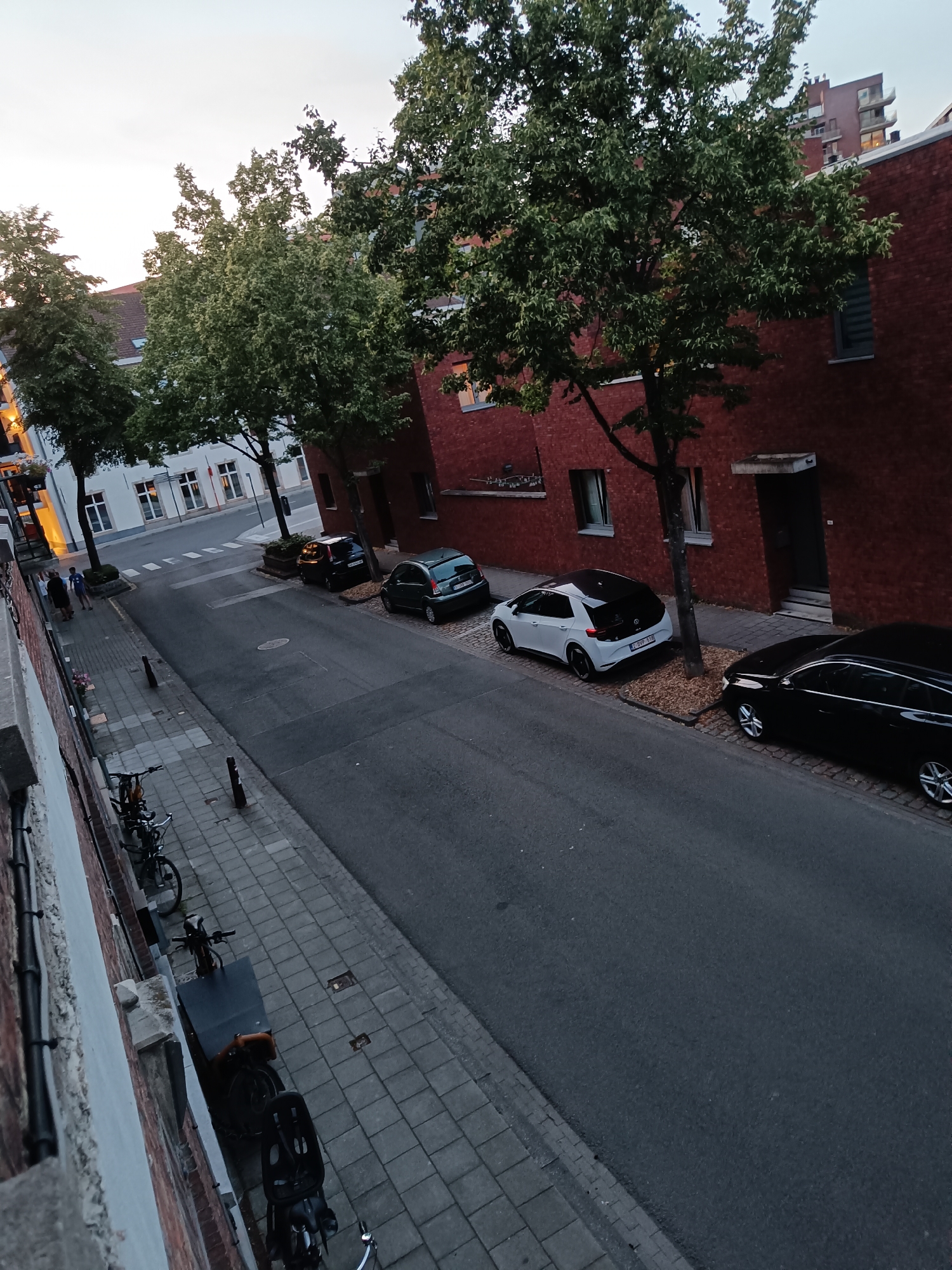 The image shows a street scene in the city of Leuven, Belgium. The street is paved with asphalt and lined with sidewalks. Several cars are parked on the street and sidewalk, including a black sedan, a white hatchback with a black roof, and a dark colored SUV. There are also several bicycles parked along the sidewalk. Tall trees with green foliage line the street, and brick buildings with windows can be seen on one side of the street. In the background, a taller modern building with balconies is visible. A few people are walking on the sidewalk in the distance. The sky is overcast, suggesting it is either early morning or late evening. The scene appears to be a quiet residential or commercial street.