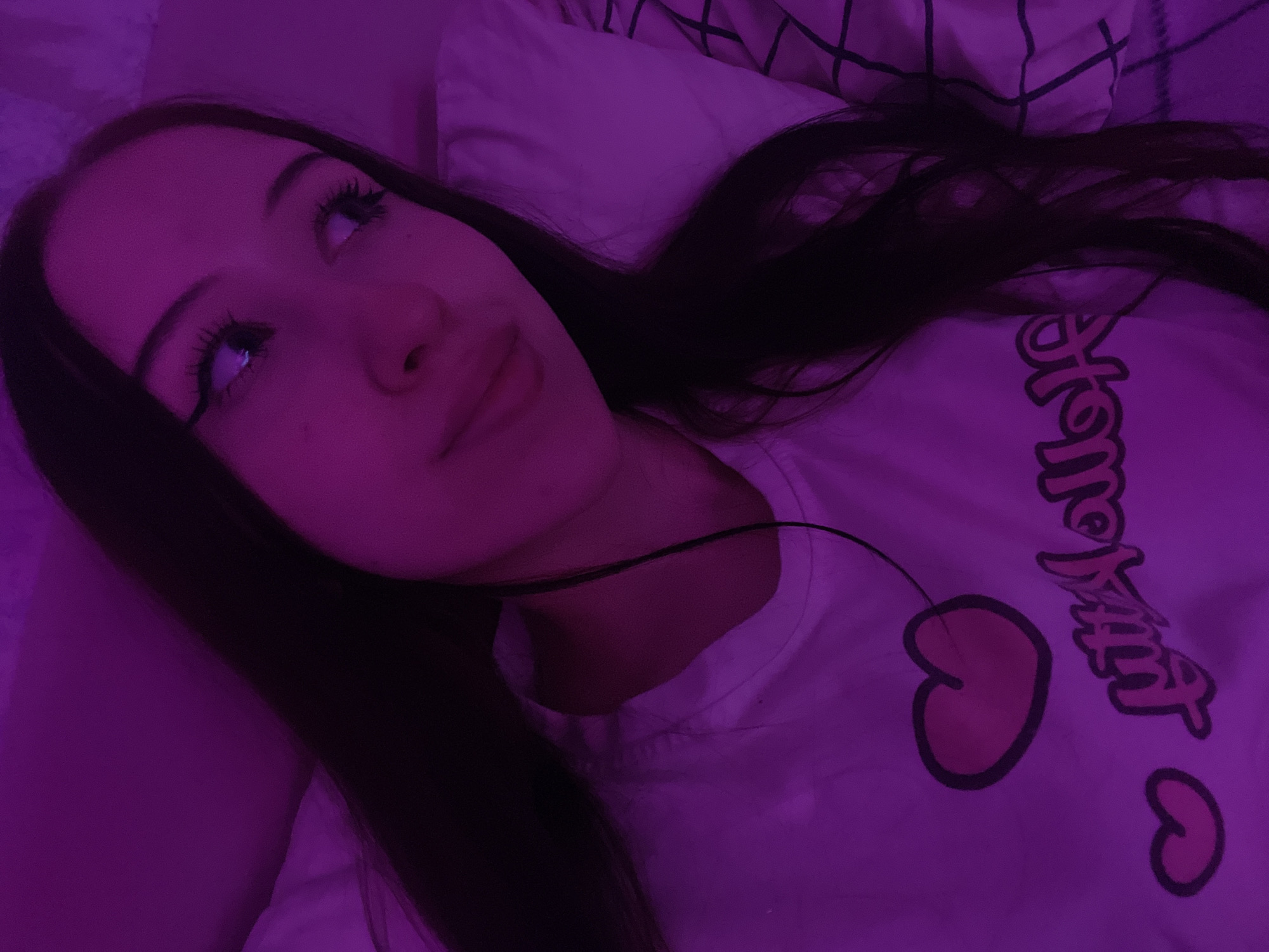 A young person with long, dark hair is lying down, looking up and to the left with a thoughtful expression. They are wearing a white t-shirt with a pink graphic design featuring cherries and stylized text. The t-shirt's text appears to read "honey." The overall scene is bathed in a purple light, suggesting an artificial light source or possibly a dimly lit room with colored lighting. The background shows a white pillow with a black grid pattern, indicating an indoor setting, likely a bed. The lighting creates a soft, dreamlike atmosphere. The image appears to be a close-up portrait, focusing on the subject's face and upper body. No specific time of day or weather conditions can be determined from the image. The location context provided is Votkinsk, Russia.
