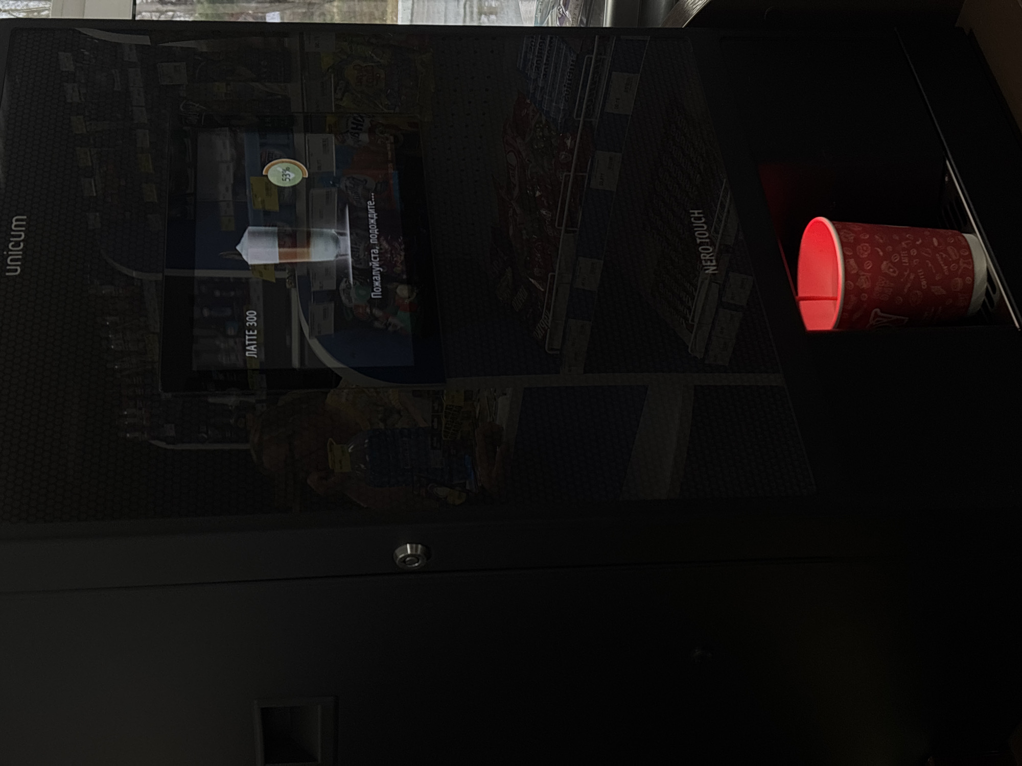 Here is a description of the image:
The image shows a Unicum Nero Touch coffee vending machine. The machine's screen displays the progress of a latte being prepared, with the text "Пожалуйста, подождите..." (Please wait...) and a progress indicator at 53%. A red and white disposable cup is positioned in the dispensing area to the right of the screen. The setting appears to be indoors, possibly in Chaikovsky, Russia, though the specific location details are sparse.
