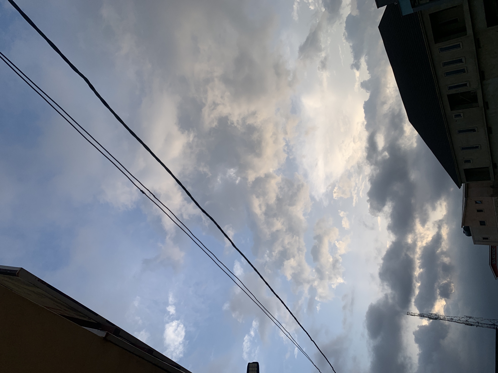 The image shows a view looking upwards in Gura Top, Nigeria. The sky is filled with a mix of light blue and grey clouds, with the sun appearing to shine through the clouds. Two black power lines are diagonally crossing the view. Sections of buildings border the image, including sections of a dark-colored roof at the bottom left and a larger building on the right. A tower is also visible between the clouds in the background.