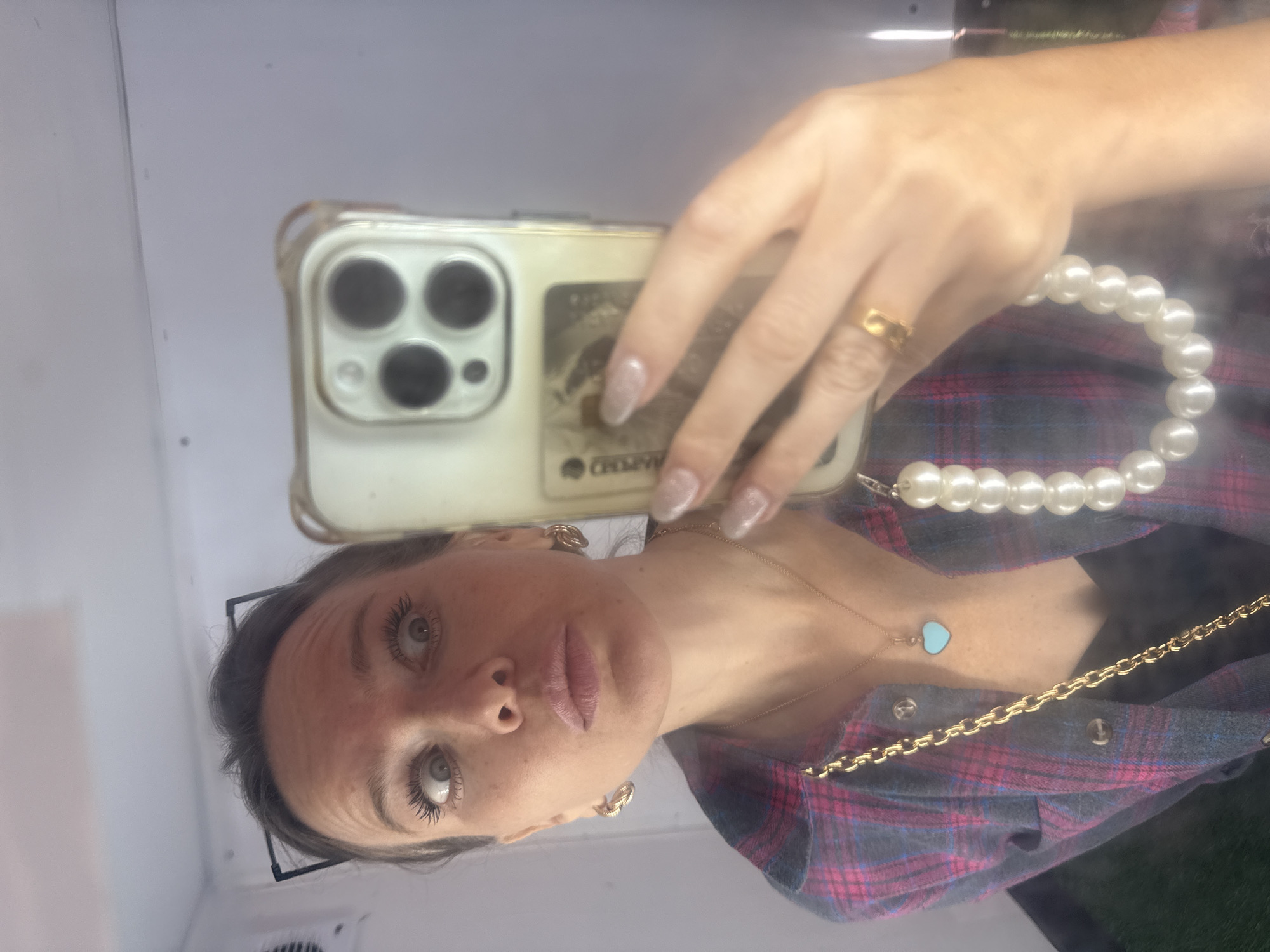 This is a selfie taken in a room with white walls, possibly a bathroom or changing room, given the reflective surface. The subject is a woman with light-colored eyes and dark hair, wearing a plaid shirt and jewelry. She is holding up a smartphone with a clear case, which has a credit card or identification card partially visible in the case behind the phone. The phone's camera is pointed towards a mirror, capturing her reflection.
The woman is wearing a gold ring on her left ring finger and a necklace with a small, light blue heart-shaped pendant. She also has hoop earrings and a string of pearls draped around her neck, with one end resting on her shoulder. Her fingernails appear to be manicured and possibly have a glittery finish. Her expression is neutral, with her eyes looking directly at the camera.
The image is vertically oriented and taken from a low angle, looking up at the subject. The lighting appears to be artificial, and there are some minor distortions and blurs, common in mirror selfies. There is no indication of the time of day or weather outside. The setting suggests an indoor, private space. No visible text other than what is on the card in the phone case, which is partially obscured and unreadable. The context of Moscow, Russia, is not visually evident in the image itself.