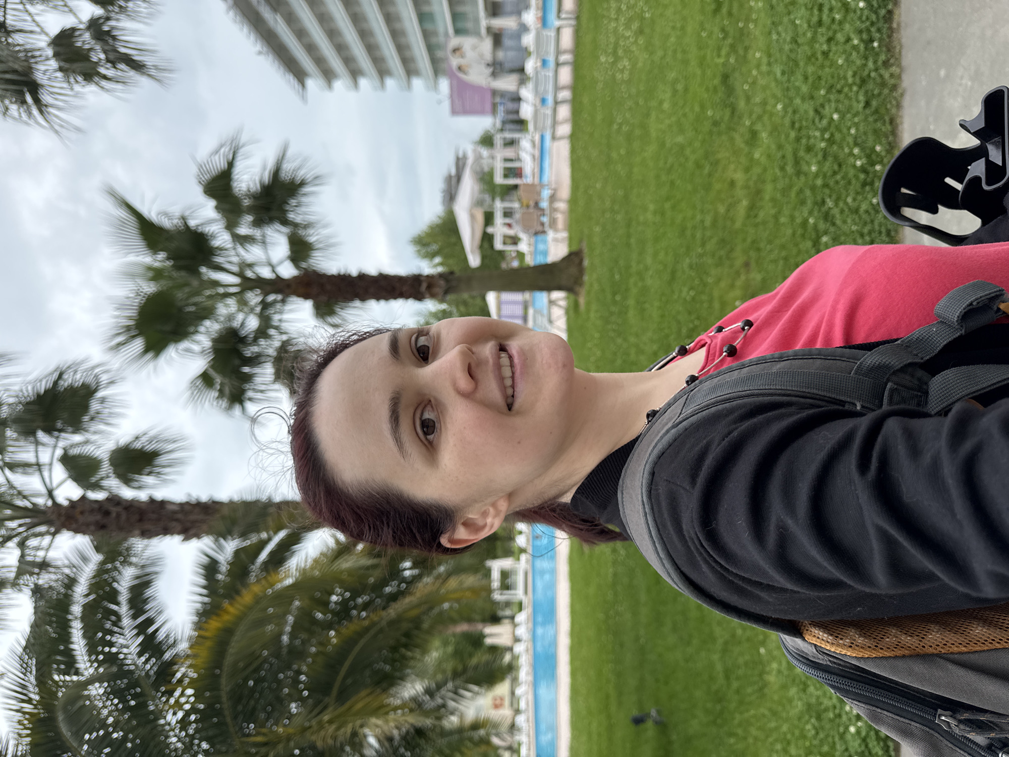 A woman is shown from the chest up, smiling and looking slightly up and to the left. She has fair skin, dark brown hair, and is wearing a red top under a black jacket, with a black backpack on her back. Behind her, palm trees line the edge of a swimming pool with turquoise water. Beyond the pool is a green lawn, and in the background, a multi-story building is visible. The scene is likely located in Sochi, Russia, given the presence of palm trees, which are not native to most of Russia but are commonly found in Sochi's subtropical climate. The image appears to be taken outdoors on an overcast day.