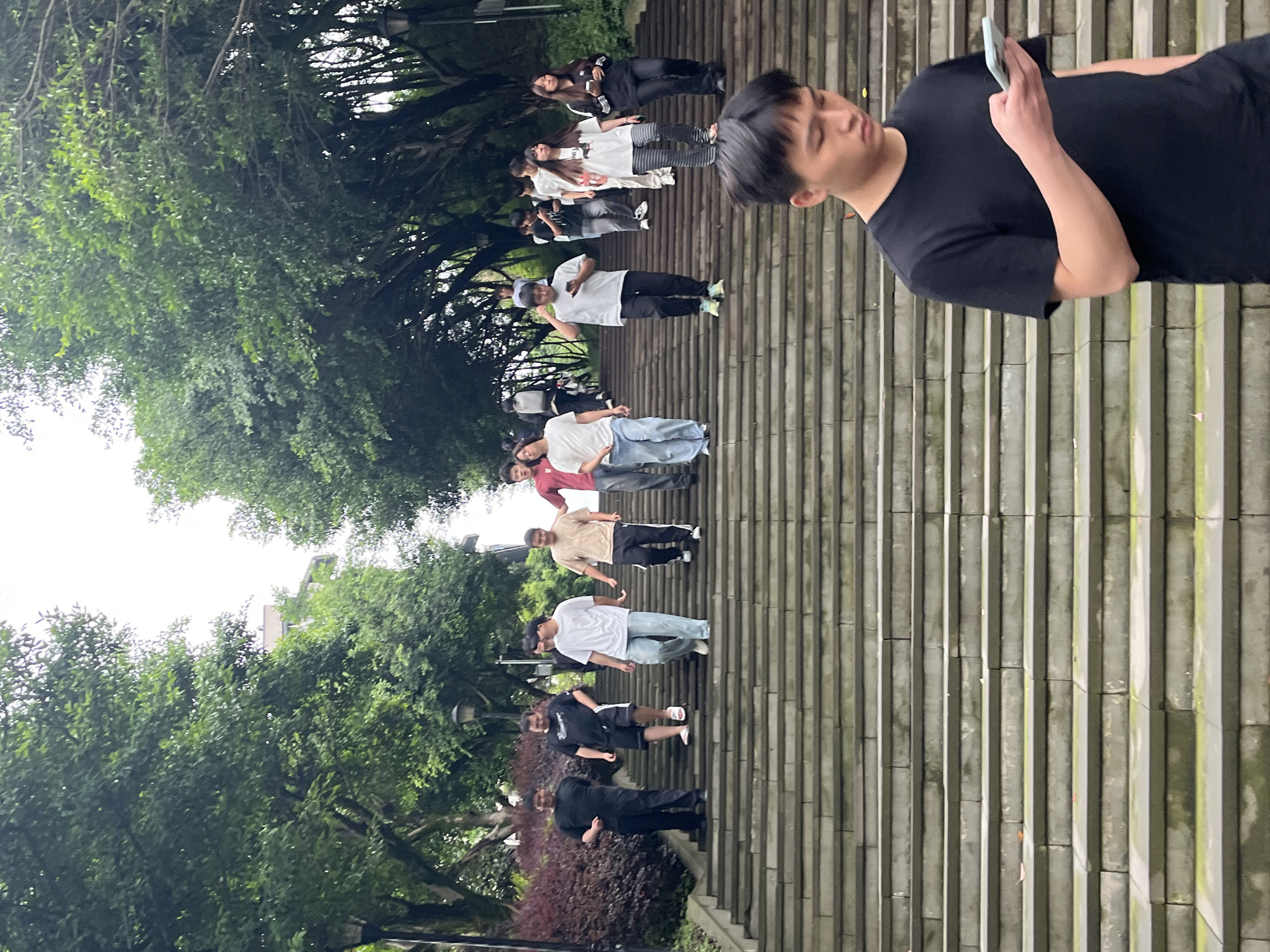 This image captures a group of young people descending a wide, outdoor stone staircase in a park-like setting with abundant green trees and foliage, likely on the campus of Chongqing University of Posts and Telecommunications in Jiangjin District, Chongqing, China. The stairs are arranged in long, evenly spaced rows. In the foreground, a young man in a black t-shirt is looking down at his smartphone. Further up the stairs, several individuals, predominantly young adults, are walking downwards. They are dressed in casual attire, including t-shirts, shorts, and pants. The lighting suggests an overcast day.