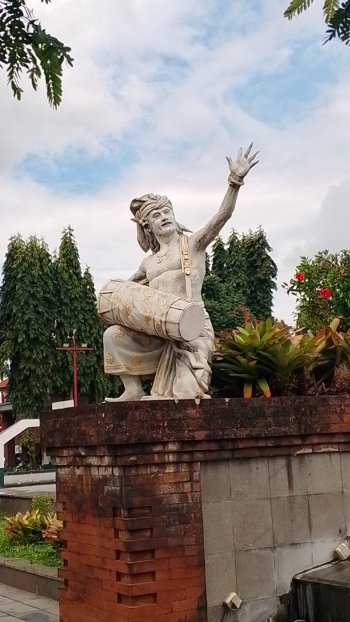 A statue of a man playing a drum is shown in front of a building. The statue is made of stone and depicts the man in traditional clothing, with a turban on his head and a sarong around his waist. He is holding a drum in his lap and playing it with his hands. The statue is placed on a stone pedestal in front of a brick wall. In the background, there are trees and a cloudy sky. The scene appears to be outdoors, possibly in a public park or square.
