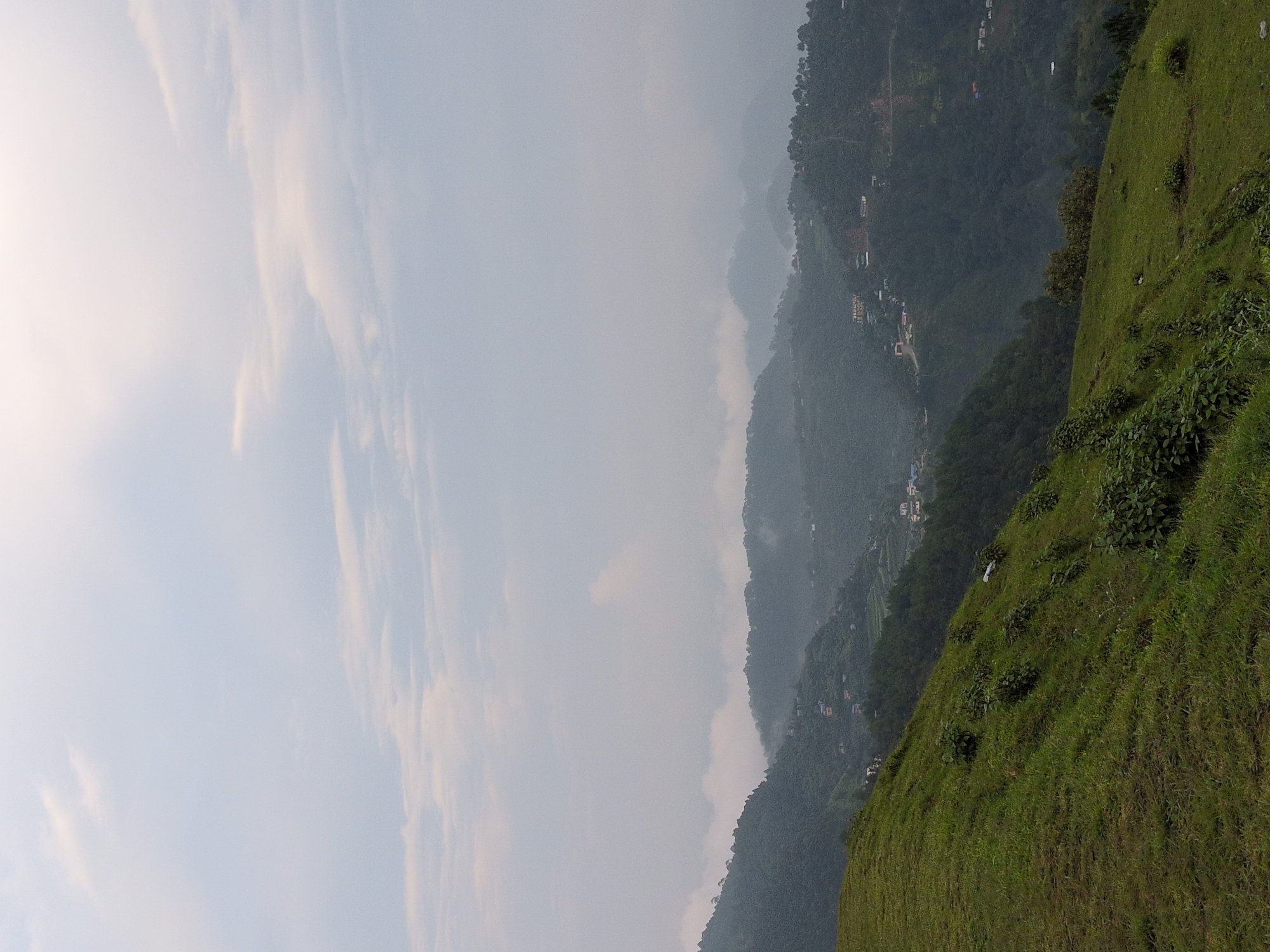 The image depicts a panoramic view of a lush, green mountainside in Kapurkot, Nepal. In the foreground, a gently sloping hill covered in dense green grass and scattered shrubs dominates the left side of the frame. The grass appears well-maintained, suggesting it could be a park or a natural reserve.
Beyond the foreground hill, the landscape unfolds into a series of rolling, forested mountains that recede into the distance. The mountains are covered with a thick canopy of trees, creating a rich, dark green texture. The terrain is uneven, with steep slopes and valleys.
Scattered across the hillsides are small clusters of buildings, indicating human habitation. These appear to be houses and possibly some small businesses, nestled into the natural contours of the land. Roads are visible winding through the mountains, connecting these settlements.
The sky above is overcast with a layer of soft, diffused clouds. The light is muted, giving the scene a serene and somewhat hazy atmosphere, characteristic of a cloudy day in a mountainous region. There are no people or animals visible in the image. The overall impression is one of natural beauty and rural tranquility.