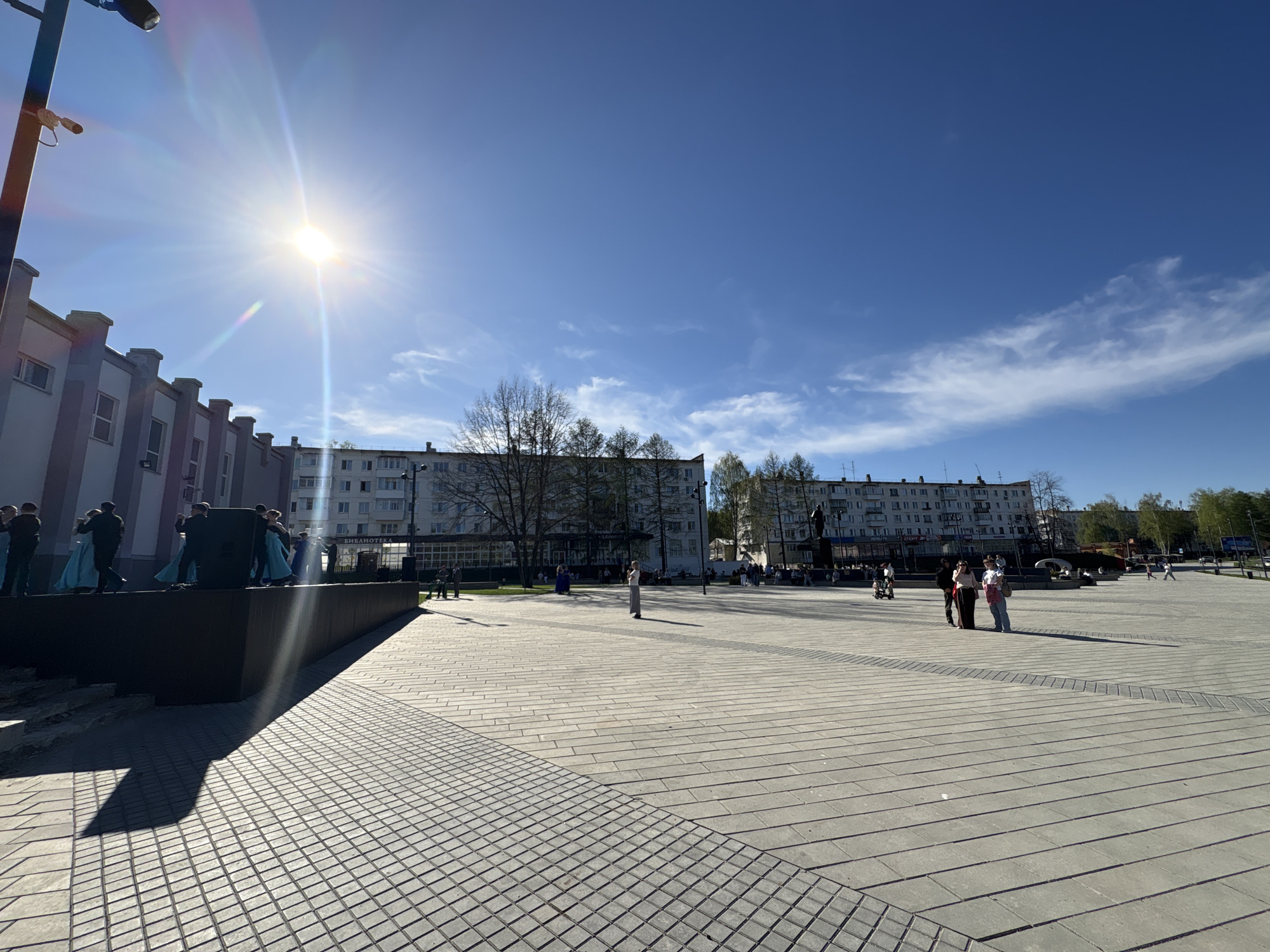 The image captures a sunny day in what appears to be a town square in Chaikovsky, Russia. The square is paved with rectangular and square paving stones. In the background are multi-story apartment buildings typical of Russian urban architecture. To the left is a modern building with a raised platform where people are gathered, some dressed in what appear to be performance costumes (light blue dresses). The sun is high in the sky, creating a bright glare and long shadows on the pavement. Several people are visible in the square, standing or walking. A light pole is present on the left of the frame. The overall atmosphere is bright and open, suggesting an urban public space in use.