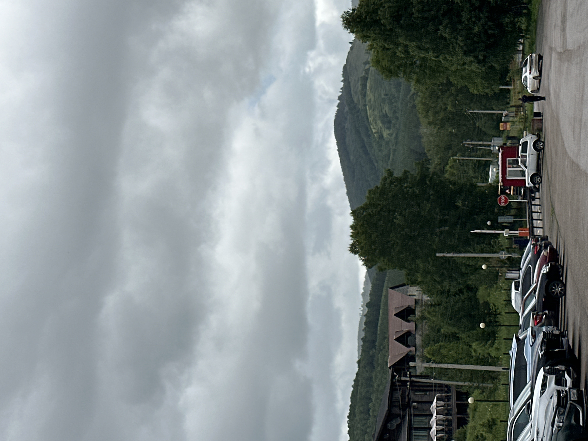The image depicts an outdoor scene with a road lined with parked cars. In the background, a lush green hillside is covered with dense trees, reaching towards a cloudy sky. The clouds are heavy and gray, suggesting an overcast or potentially rainy day. A few scattered streetlights are visible along the side of the road, some with spherical lights.
Several cars are parked in a row along the right side of the road. A white car is parked further up the road, and a person in dark clothing stands near it. Closer to the foreground, there's a red structure, possibly a small building or a kiosk, with a white SUV parked next to it. A red stop sign is prominently displayed on a pole near the red structure. Further down the road, more cars are parked, including several dark-colored SUVs and sedans.
To the left of the road, there are more trees and a glimpse of some buildings with distinctive triangular roofs. The overall setting appears to be a somewhat rural or scenic area, perhaps a mountain resort or a quiet road in a hilly region. The time of day is not clearly indicated but the lighting suggests it could be daytime under heavy cloud cover. The mood is subdued, likely due to the overcast weather. No discernible text other than the stop sign is visible.