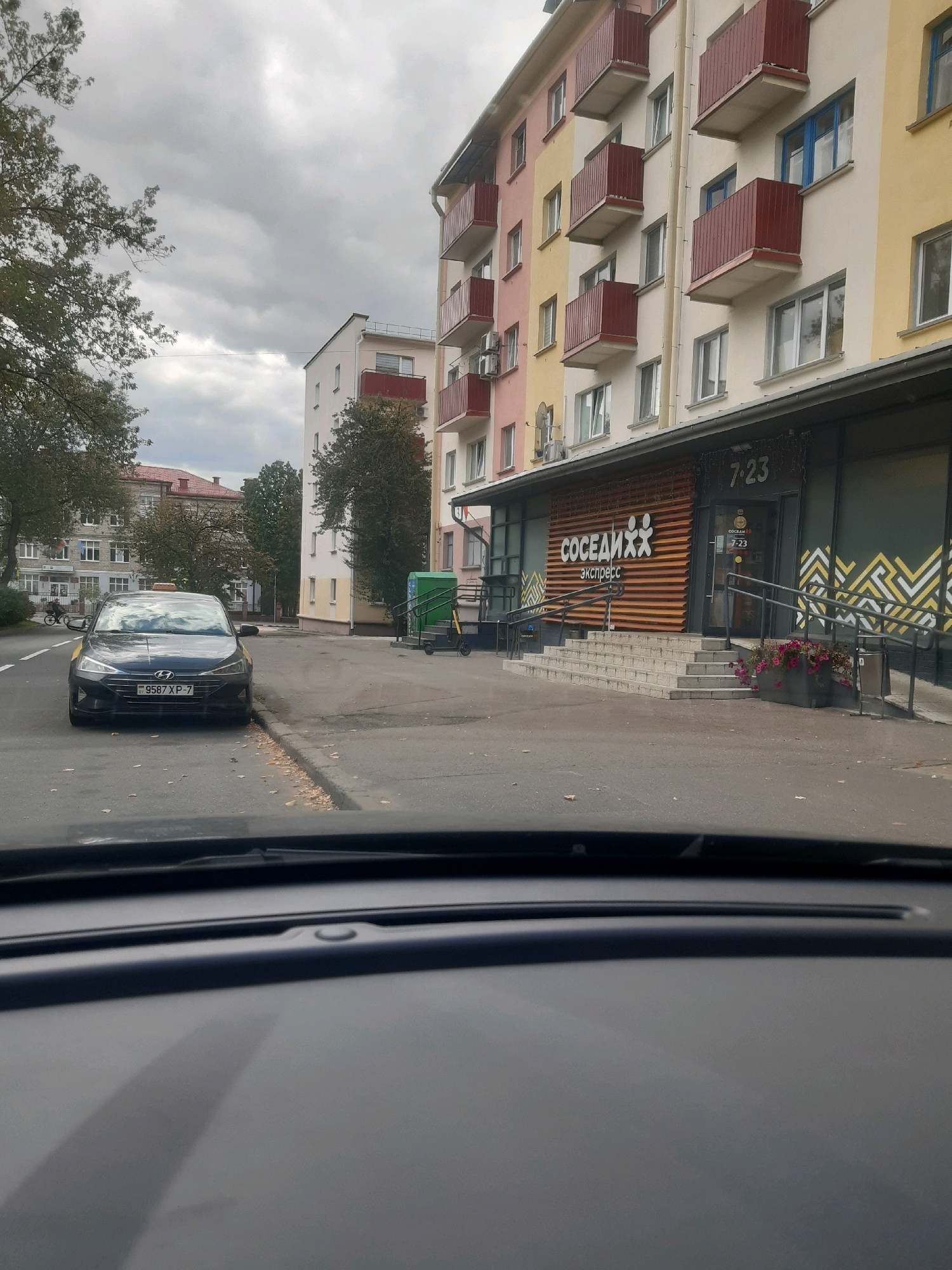 This image depicts a street scene in Minsk, Belarus, focused on a modern apartment building with a ground-floor commercial space. A dark-colored Hyundai sedan, presumably a taxi based on the yellow markings, is parked curbside on the left. The apartment building features colorful facades in shades of pink, yellow, and white, with red-railed balconies. The commercial space below has a wooden slat design on its facade, with a prominent sign for "СОСЕДИИХ экспресс" (Sosedykh Express) and the operating hours "7-23" visible. A set of stairs leads up to the entrance, with ramps on either side. In the background, other buildings and trees are visible under an overcast sky. The image is taken from the perspective of a car interior, showing the dashboard in the foreground.