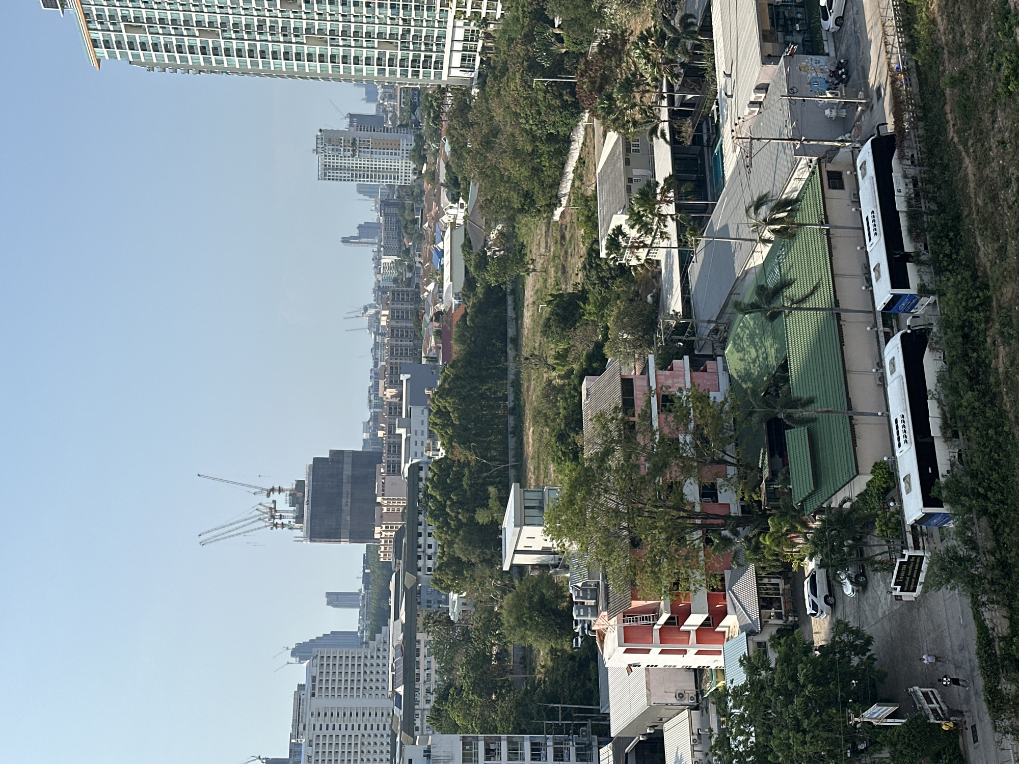 The original image is rotated 90 degrees counter-clockwise. Describing it as if correctly oriented (landscape), it presents an expansive aerial view of a vibrant cityscape, likely Pattaya City, Thailand, based on the provided context.
The scene captures a mix of urban development and natural elements under a clear, bright blue sky, indicating daytime. In the foreground, a street or parking area runs along the right side, where several large, white tour buses are parked. One bus clearly shows "CITY TOUR" and "CLEAN BUSES" on its side, suggesting a tourism focus. A white car and a motorcycle are also visible, along with a couple of people walking on the paved surface. To the left of the street are various lower-rise buildings with colorful roofs (green, red, pink, white), interspersed with lush green trees, including prominent palm trees. A small white sign with black Thai script is visible near the street but is not clearly legible.
Moving towards the mid-ground, a wide strip of undeveloped, overgrown land runs horizontally across the image, separating the foreground buildings from more distant structures. Beyond this green space, the cityscape extends into the distance, dominated by numerous high-rise apartment or condominium buildings. Several large tower cranes are visible, especially in the mid-ground and further left, indicating ongoing construction and urban expansion. The overall impression is one of a bustling, developing coastal city with a blend of residential, commercial, and natural areas. The weather appears sunny and clear.