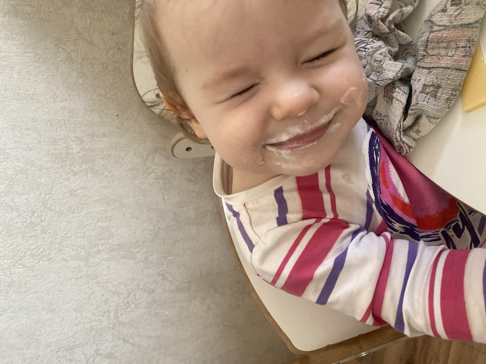 A close-up, slightly high-angle shot captures a baby in what appears to be a high chair, likely during or after a meal. The baby's face is central to the image, covered in a white, creamy substance around the mouth, nose, and cheeks, suggesting food like yogurt or a mashed meal. The baby has light-colored hair and their eyes are closed, indicating a strong expression, possibly of enjoyment, a sneeze, or intense concentration. A small, contented smile is visible despite the food on their face.
The baby is wearing a long-sleeved top with bold, irregular stripes in white, hot pink, and purple. A patterned fabric, possibly a bib or blanket with geometric designs in muted colors, is draped over the baby's right shoulder and chest area. To the far right, on a white tray or table surface, a small, irregularly shaped piece of yellow food, possibly cheese, is visible.
The background to the left consists of a light, textured wall or surface. The overall impression is one of a joyful and messy moment during a baby's mealtime. No text is visible in the image.
