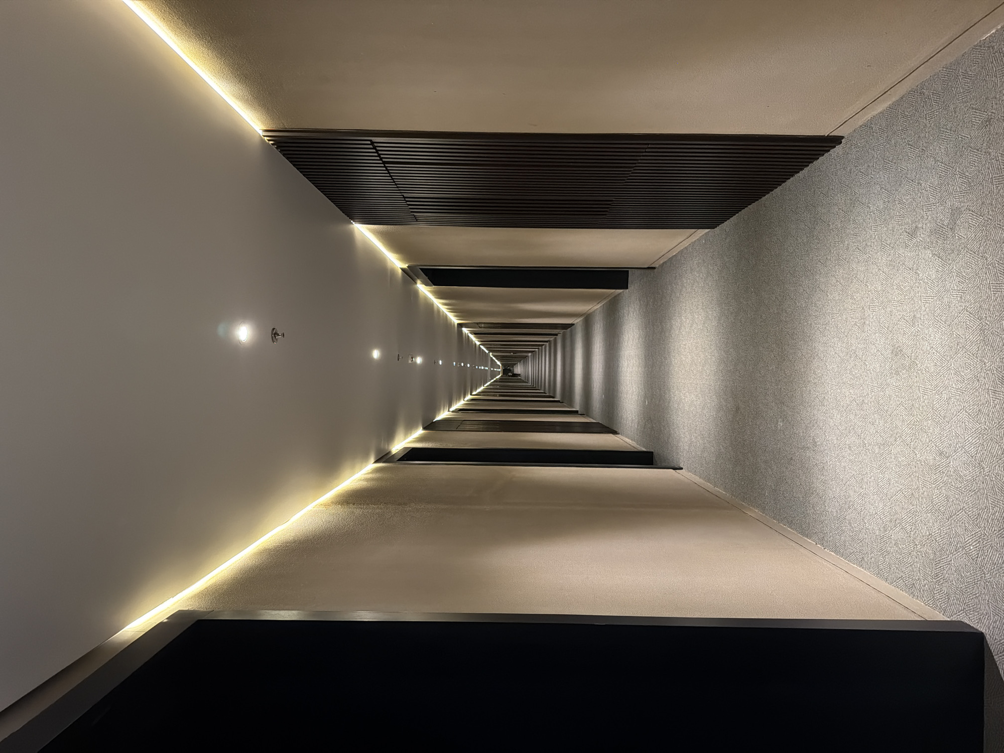The image captures a long, empty indoor corridor, seen from a low perspective, extending into the distance to a clear vanishing point. The overall aesthetic is modern and sophisticated, characterized by clean lines and thoughtful lighting.
The left wall of the hallway is a light, smooth surface, possibly beige or off-white. Near its junction with the ceiling, a dark, slatted decorative panel runs horizontally along the corridor's length. The right wall is covered in a light grey carpet with a subtle, repeating geometric pattern.
The ceiling is plain and light-colored. It features linear LED strip lighting running along its edge with the left wall, casting a warm yellow glow. Several recessed spotlights are also visible on the ceiling at regular intervals. A small fire sprinkler head is present on the ceiling in the foreground.
The floor is marked by a series of dark, rectangular, raised panels that extend down the hallway. Strip lighting is also visible along the junction of the floor and the right wall.
No people are visible, and no specific activity is taking place in the corridor. The strong linear perspective, combined with the interplay of warm ambient strip lighting and cooler spotlights, creates a sense of depth and serene elegance. The contrasting textures of the smooth wall, slatted panel, and patterned carpet add visual interest to the minimalist design. The setting is an indoor space, likely within a hotel or contemporary building in North Kuta, Indonesia. There is no visible text.