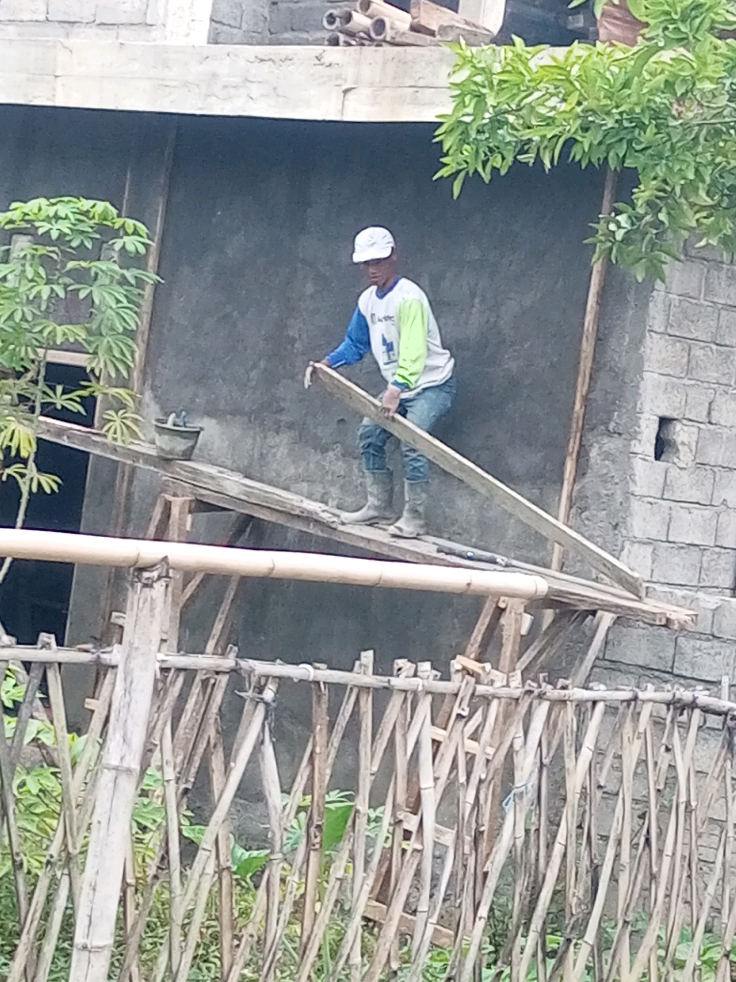 A construction worker, wearing a white hard hat and a long-sleeved shirt with blue and lime green accents, is balancing on a wooden plank. The worker is holding another plank, appearing to be in the process of construction or repair. The scene is outdoors, with a grey concrete wall and a brick wall visible in the background. Green foliage and bamboo structures are present, suggesting a rural or semi-urban environment. The image quality is low, with a blurry and pixelated appearance. The overall impression is of active construction work in a non-urbanized area.