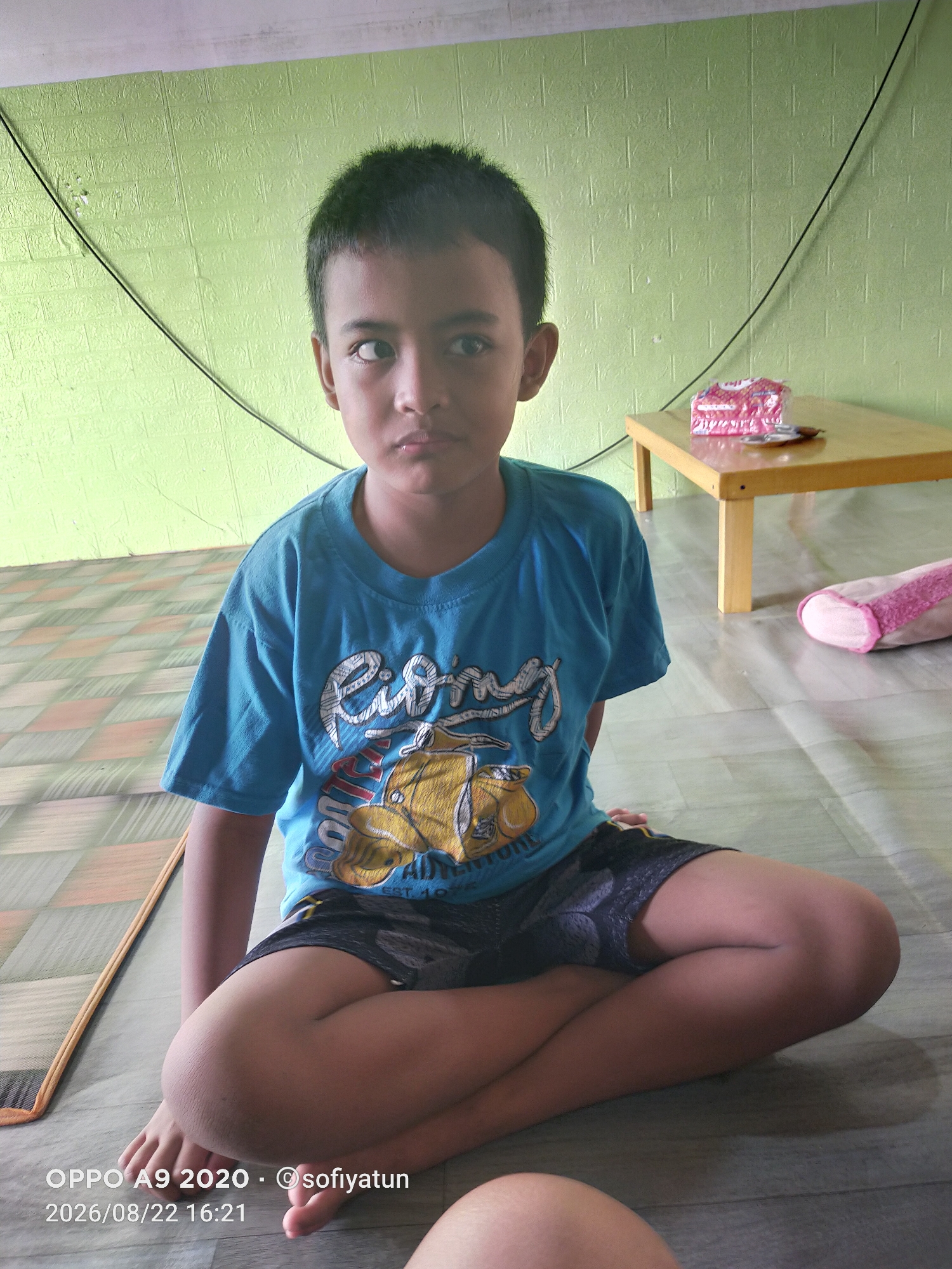 A young boy is sitting on the floor indoors, looking towards the camera with a slightly cross-eyed gaze. He is wearing a blue t-shirt with a yellow car graphic and black shorts. The room has a light green textured wall and a tiled floor. A small wooden table with a package of biscuits on it is visible in the background. A rolled-up pink and white object is on the floor to the right. The lighting suggests an indoor setting during the day. The timestamp "2026/08/22 16:21" and the text "OPPO A9 2020 - @sofiyayun" are overlaid on the image.