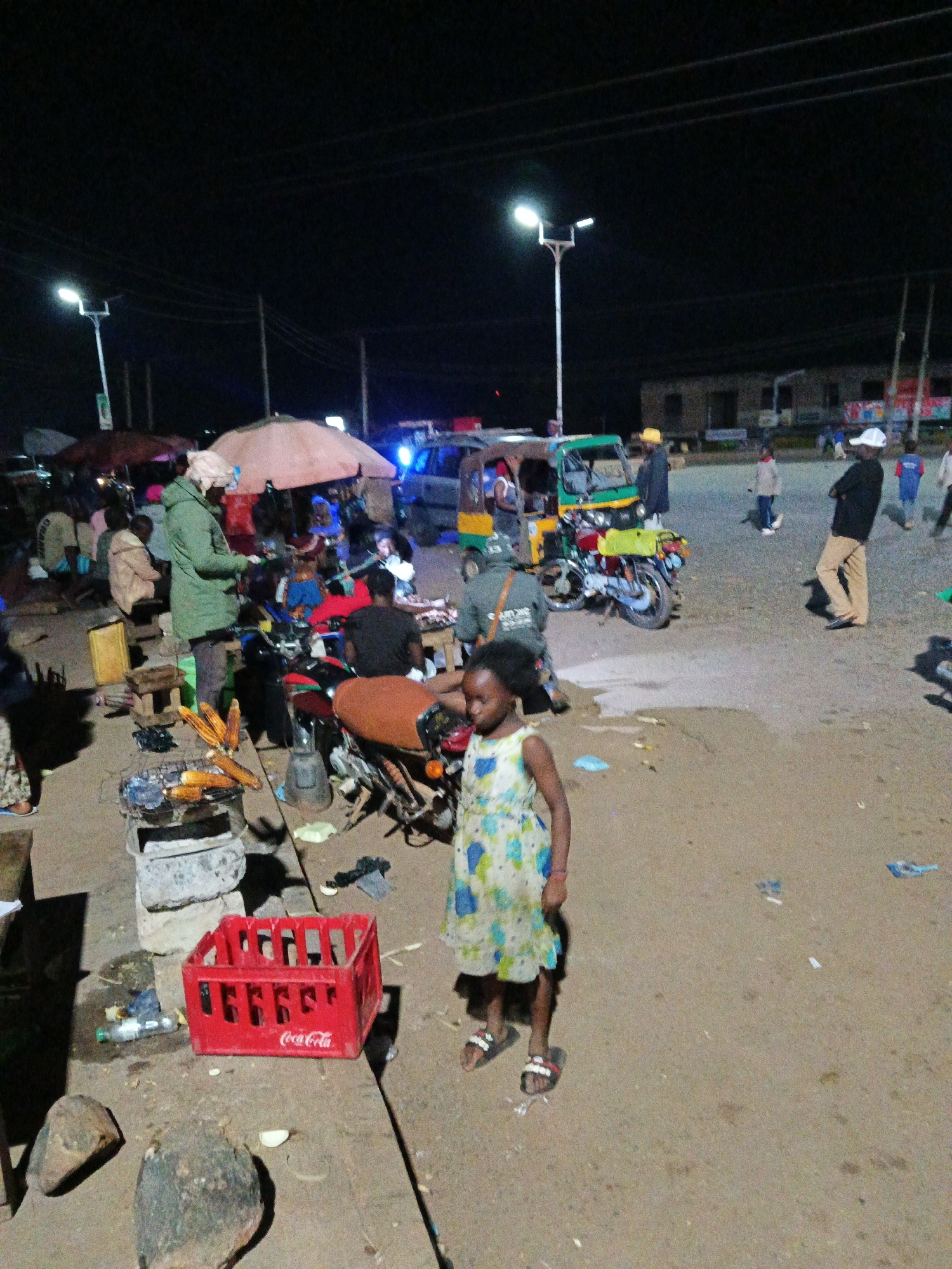 An outdoor night scene depicts a marketplace or street area with several people, vehicles, and market stalls. In the foreground, a young girl in a floral dress and sandals walks across a dusty ground. To her left is a red Coca-Cola crate. Further back, a charcoal grill is set up with corn cobs cooking. Various people are visible around market stalls, some appearing to be vendors and customers. A green and yellow tuk-tuk is parked near a motorcycle. Other people are seen in the background, including individuals walking and standing. Streetlights illuminate the scene, casting shadows. The environment appears to be a busy urban outdoor space.