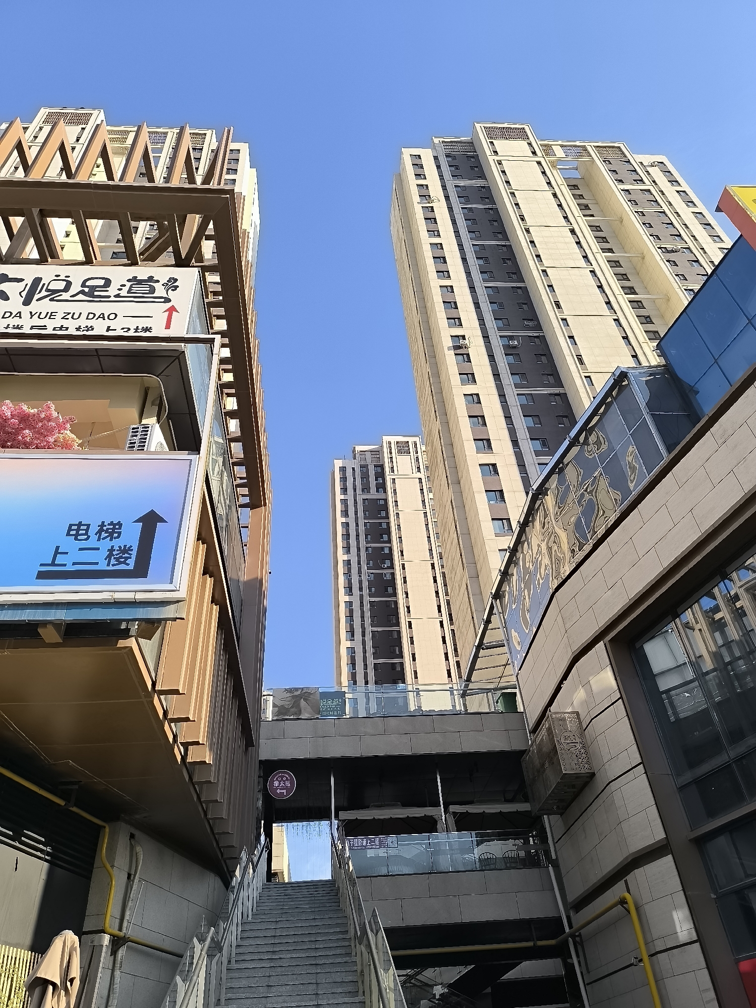 The image shows a low-angle view looking up at modern high-rise buildings and architectural elements in Ürümqi, China. The sky is clear and blue. In the foreground, there are staircases and pedestrian walkways, with metal railings and concrete structures. Signs are visible, including one with Chinese characters and an arrow pointing upwards, indicating "Elevator to the second floor." Another sign, partially obscured, appears to also be directional. The buildings are a mix of beige, white, and gray facades with numerous windows. Some structures have metallic architectural accents. The overall scene suggests an urban environment with commercial or residential complexes.
