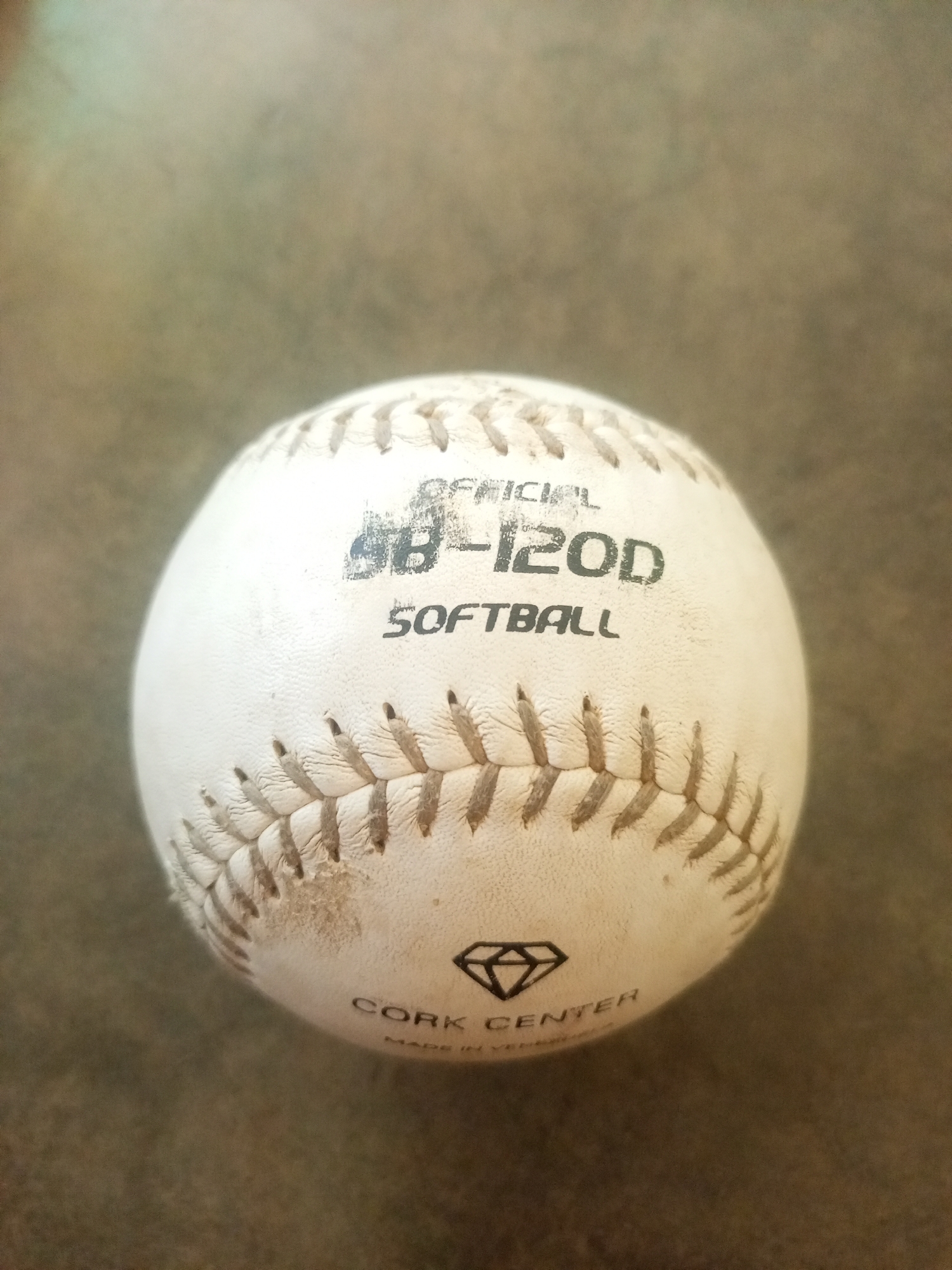 A close-up, slightly angled shot shows a white softball resting on a brown, textured surface. The softball is slightly worn and shows the characteristic red stitching. Text on the ball reads "OFFICIAL SB-120D SOFTBALL" and "CORK CENTER" with a diamond logo above it. Faint text below "CORK CENTER" appears to say "MADE IN VENEZUELA". The background is softly blurred, with muted green and brown tones. No people are present in the image. The scene is not identifiable as being in the city of La Tagua, Venezuela.